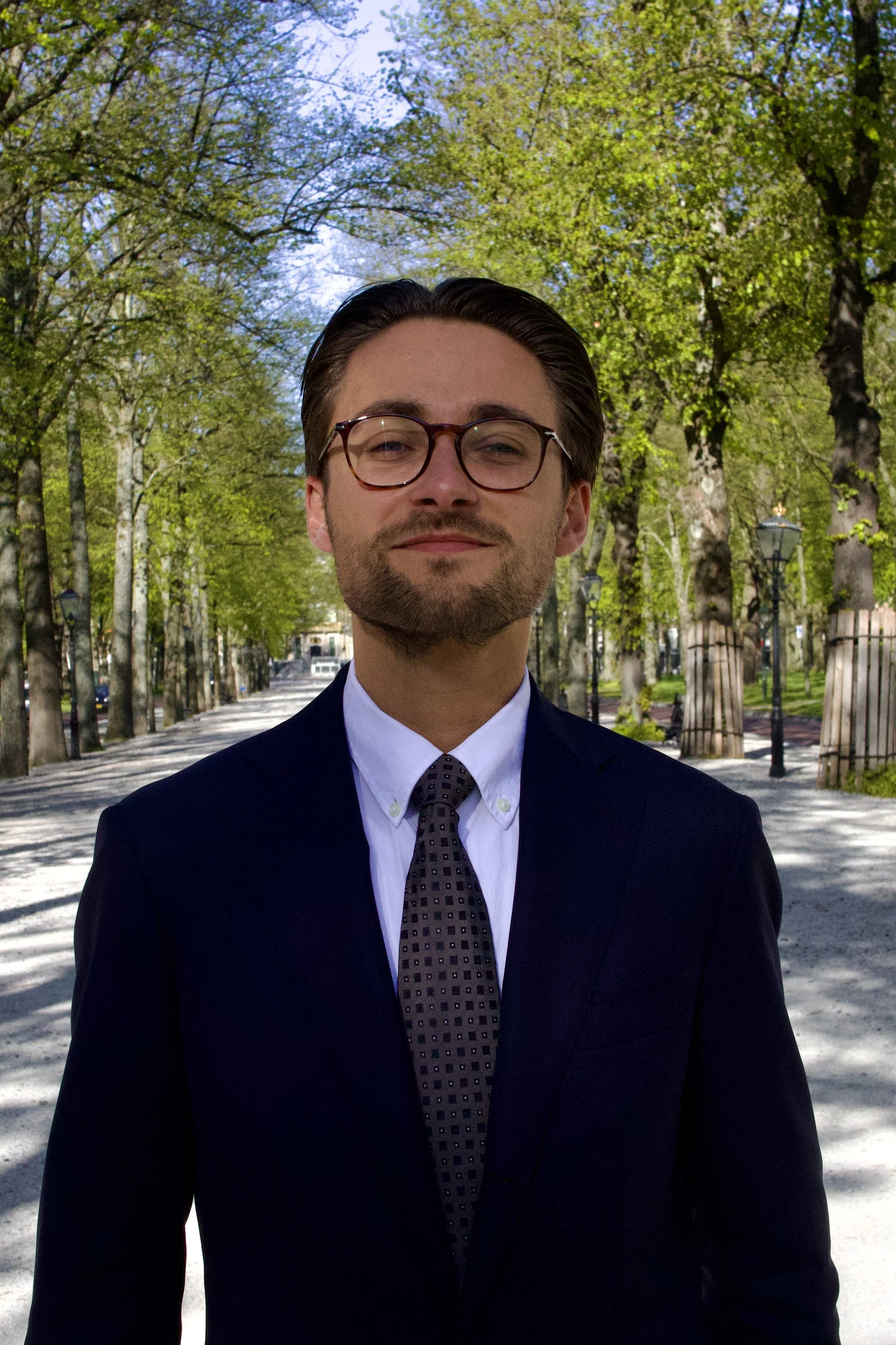 Een man in formele kleding en een bril, wandelend door een park met groene bomen en straatlampen.