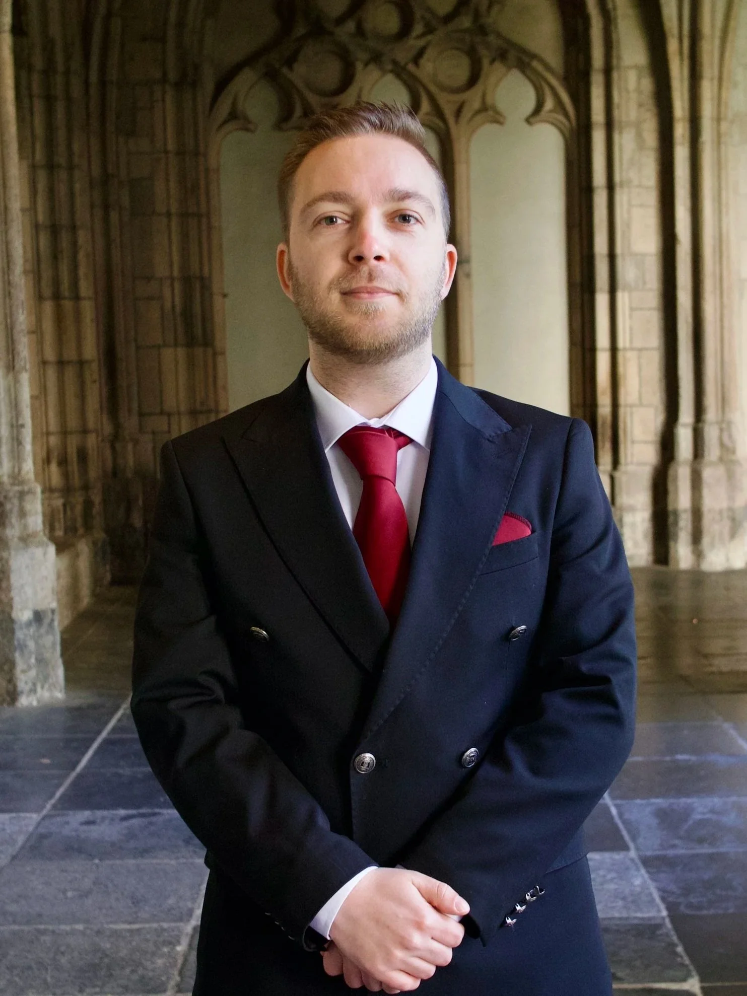 Man in formeel donkerblauw pak met rode stropdas en witte overhemd, standing voor een oude stenen kerk of kasteel, binnen een arcadereconstructie.