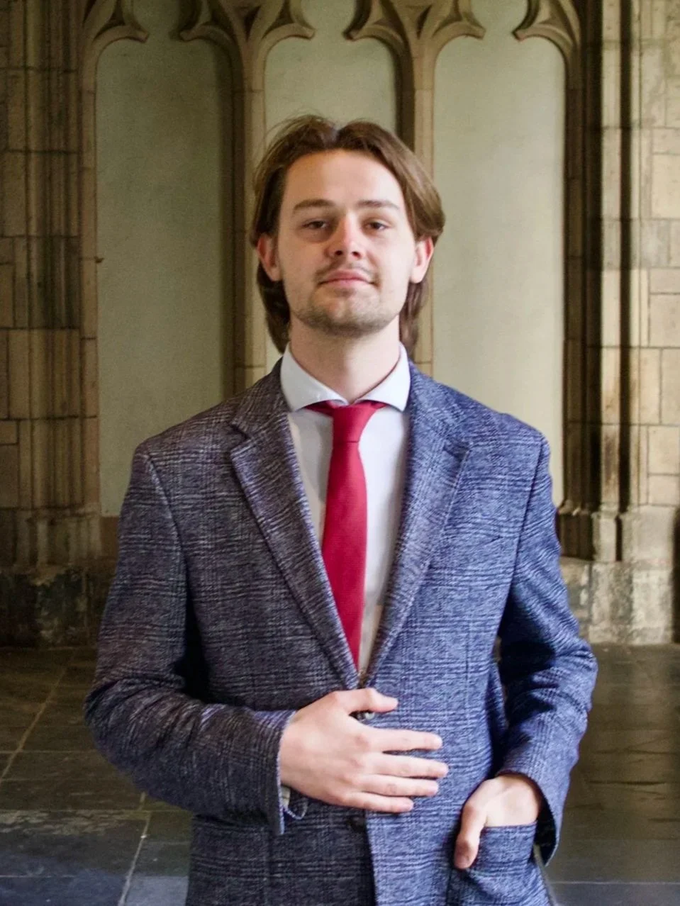 Een man in een grijs geruit pak met een witte blouse en een rode stropdas, staand binnen in eenGotische architectuur met bakstenen muren en bogen.