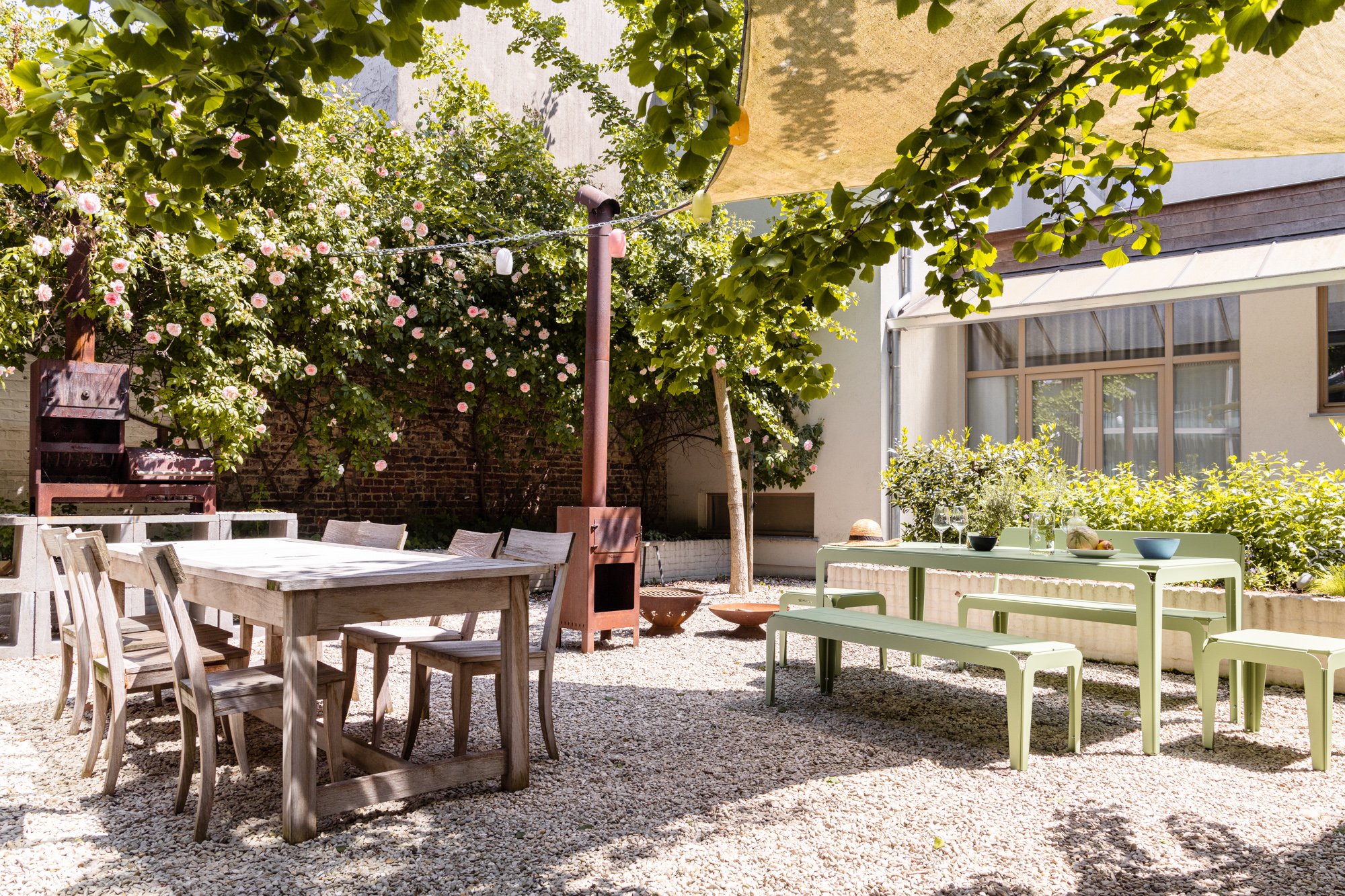 Tuin met houten tafel en stoelen, een groene tafel met banken, omgeven door bloeiende rozenstruiken en bomen, met een zonnescherm erboven.