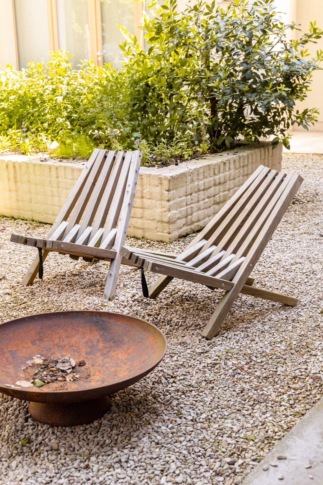 Twee houten ligstoelen op een grindterras naast een vuurkorf, omgeven door groene planten.