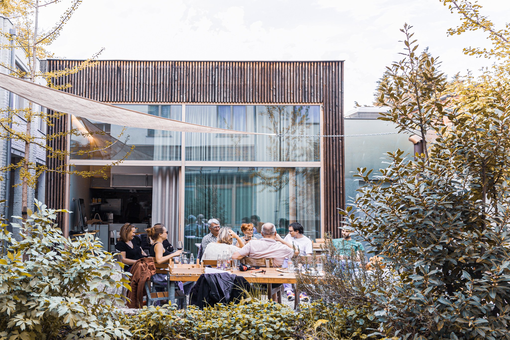 Groep mensen die buiten in een tuin om een tafel zitten, voor een modern huis met grote ramen. Omringd door planten en bomen.