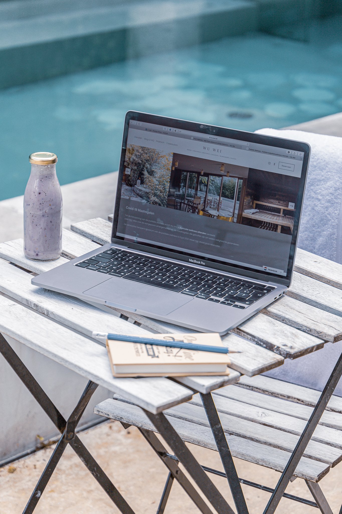Laptop op houten tafel bij zwembad, met fles drank en notitieboekje.
