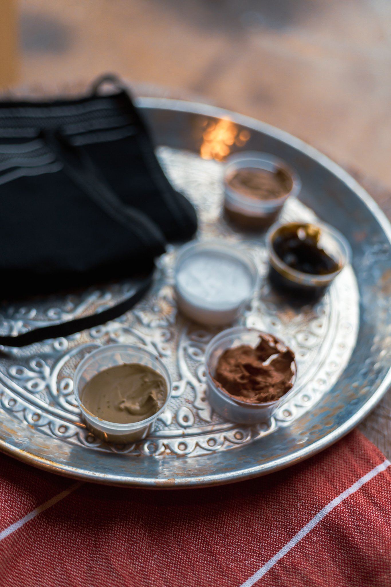 Zilveren dienblad met schaaltjes klei en zwarte doek op rode ondergrond, voor hammam scrubritueel.