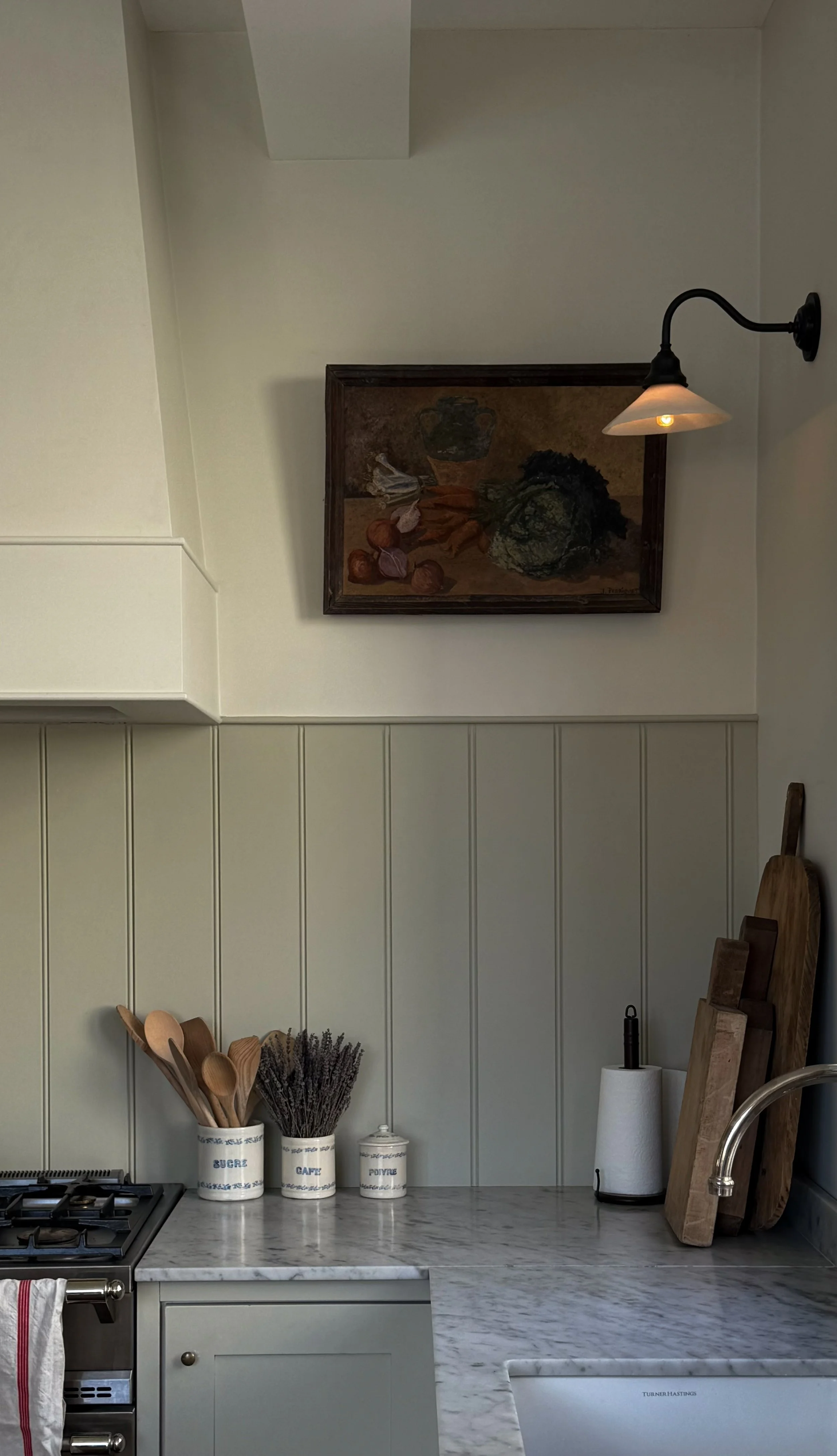 A kitchen countertop with a marble surface, wooden utensils in a ceramic jar labeled 'SUGAR' and 'COFFEE', a small jar labeled 'POIVRE' (pepper), wooden cutting boards, a paper towel roll, and a sink. Above, there is a wall-mounted lamp and a framed 