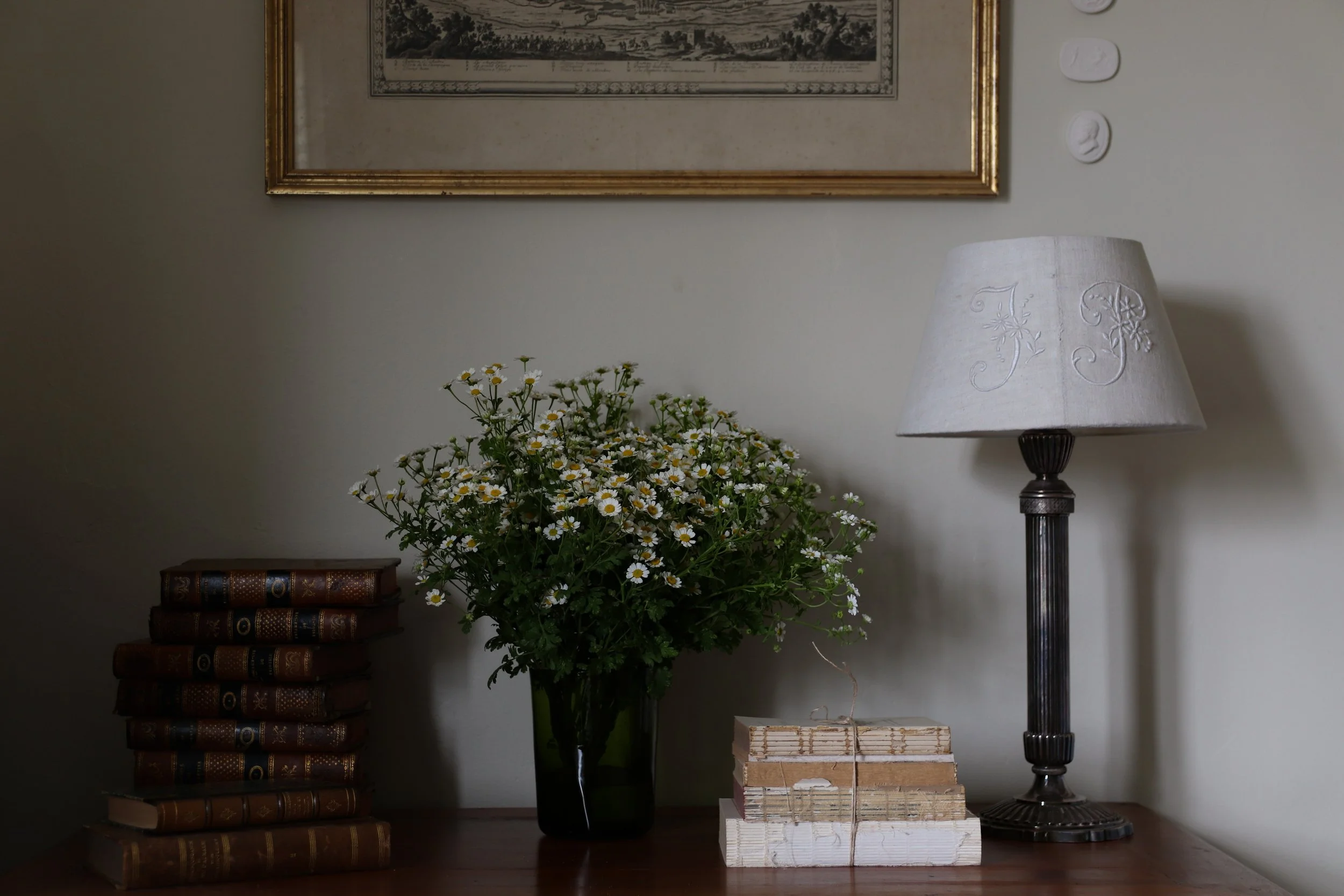 A wooden table with a stack of vintage brown books, a large green vase with white daisies, a set of wrapped beige and white paper packages, and a black lamp with a white lampshade embroidered with floral patterns. There is a framed artwork hanging on