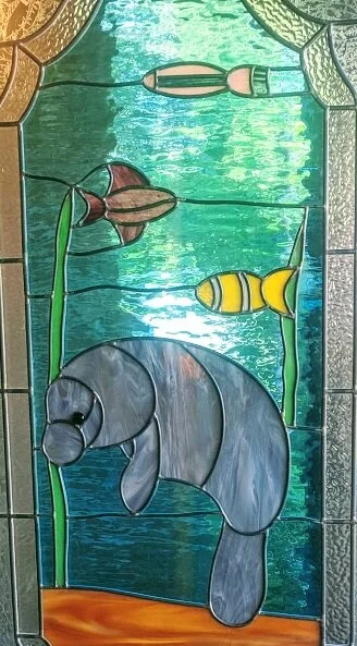 Stained glass window featuring a gray elephant, yellow fish, pink cattails, and green reeds set against a blue and green background.
