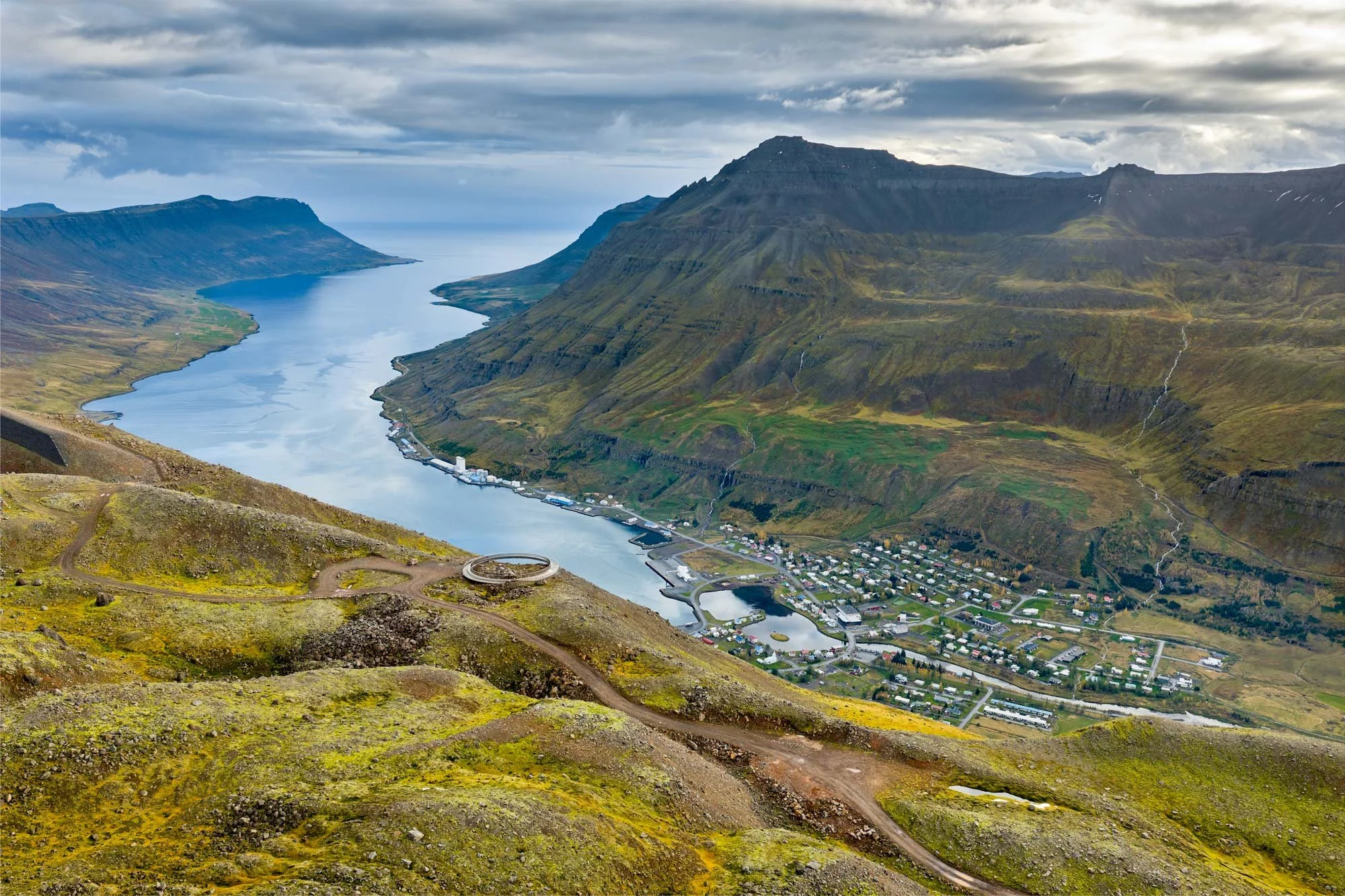 Aerial view of a fjord surrounded by steep green mountains with a small town along the shoreline and cloudy sky overhead. Baugur Bjólfs in Seyðisfjörður, East Iceland.