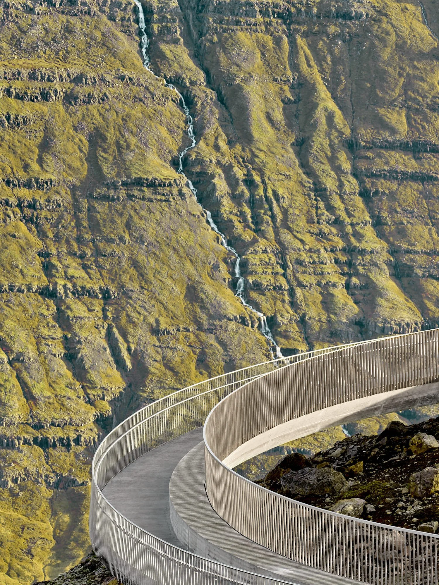 A winding walkway over a rocky terrain with a mossy mountainside and a small waterfall in the background. Baugur Bjólfs in Seyðisfjörður, East Iceland.