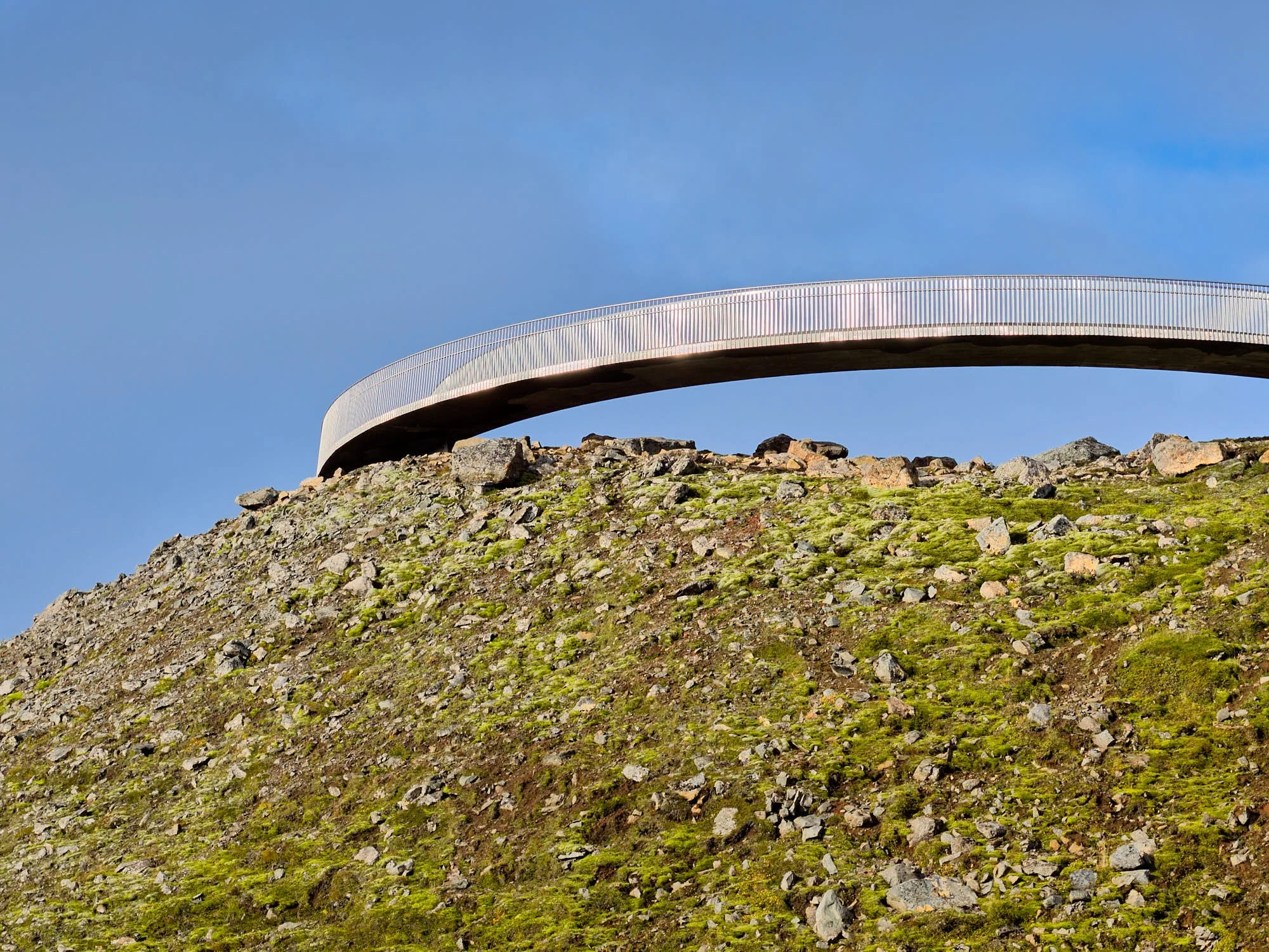 Modern curved walkway or bridge above a mossy rocky hillside, against a blue sky with some clouds. Baugur Bjólfs in Seyðisfjörður, East Iceland.