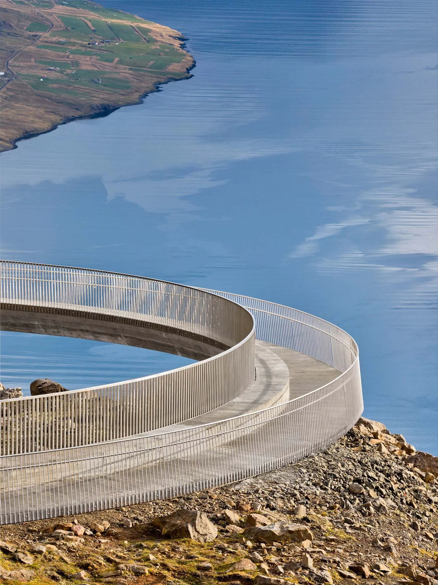 A spiral walkover bridge on a rocky hillside overlooking a large body of water reflects the blue sky and partial green land in the distance. Baugur Bjólfs in Seyðisfjörður, East Iceland.