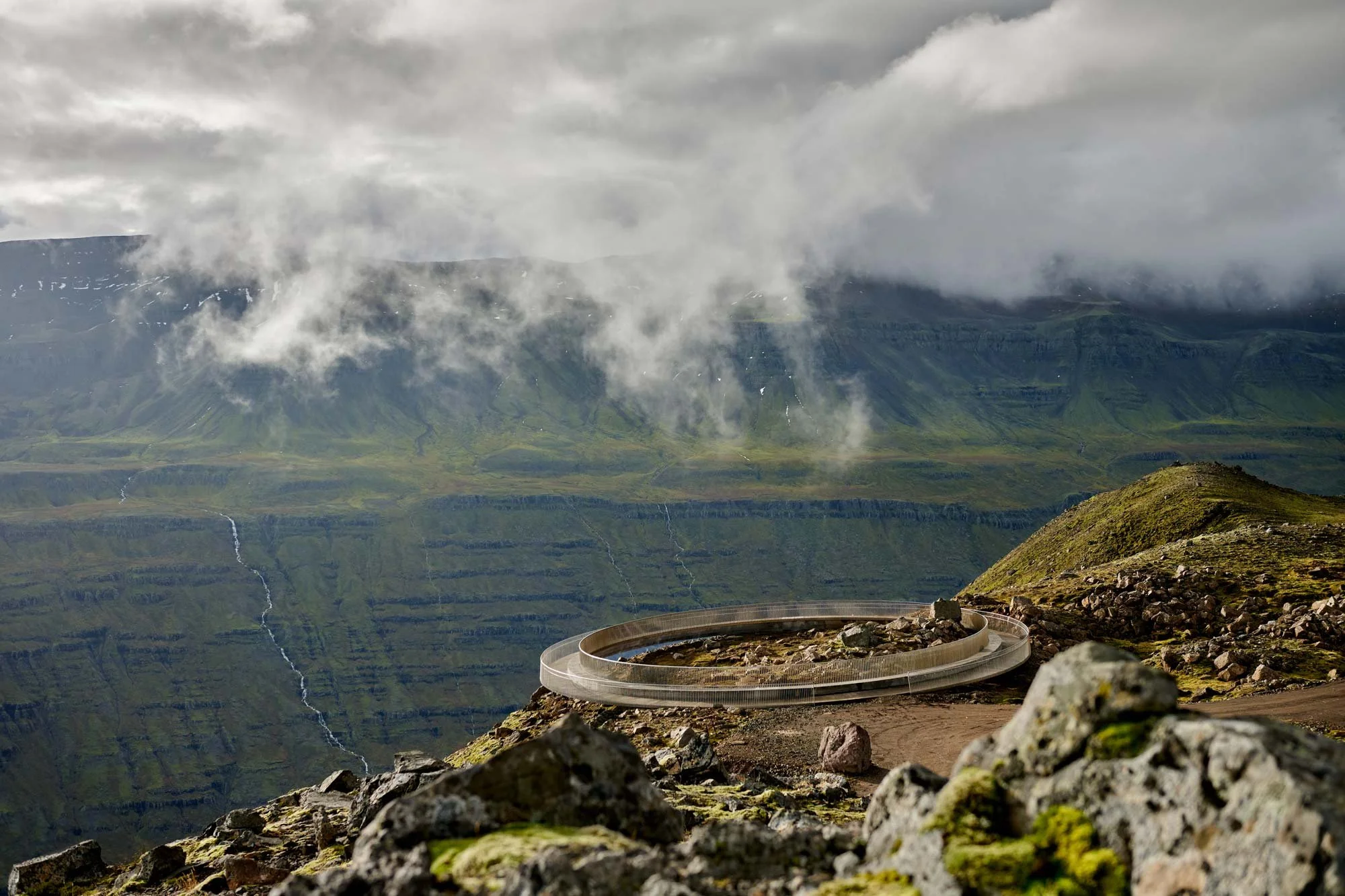 A volcano crater with a ring-shaped viewing platform in a mountainous landscape with clouds and waterfalls in the background. Baugur Bjólfs in Seyðisfjörður, East Iceland.