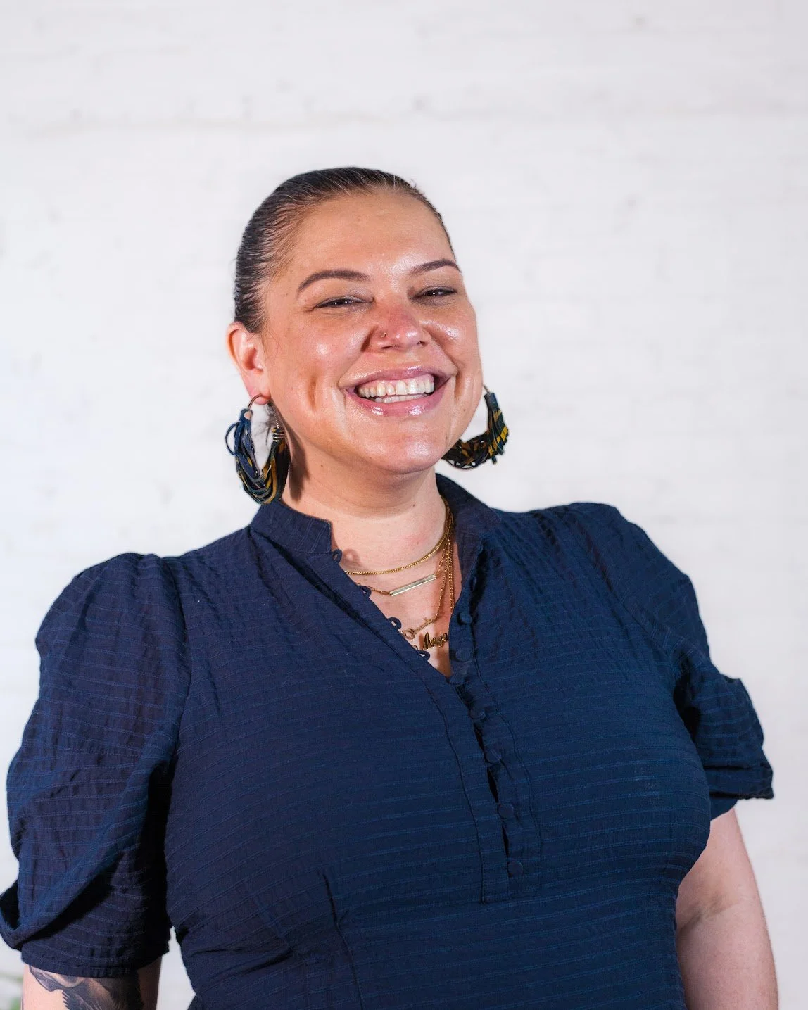 Headshot of CAF’s Executive Director Megan Jeyifo (a person with tan skin and short hair) from chest up smiling in a blue blouse and round earrings
