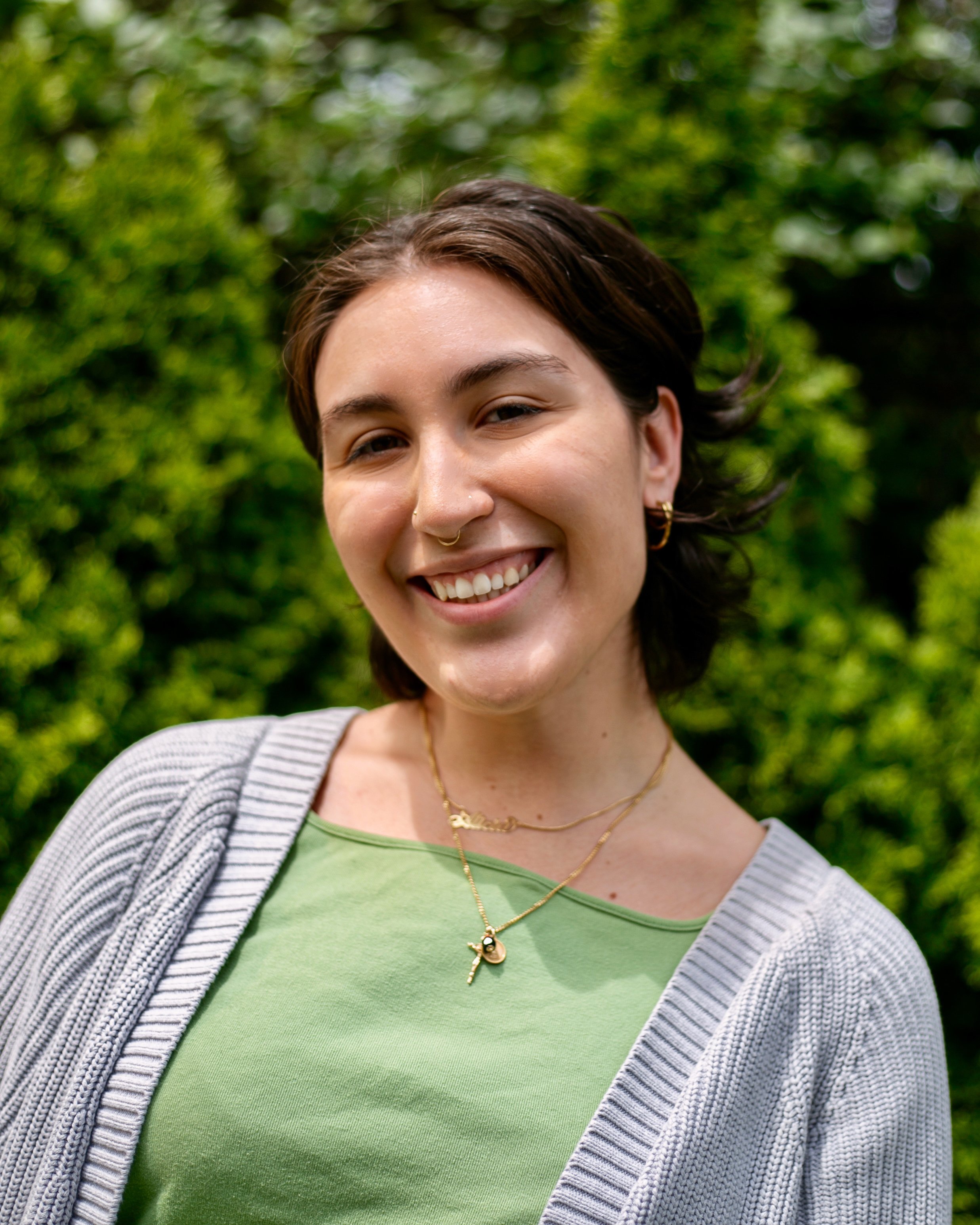 Young woman with short dark hair, wearing a green shirt with a cardigan, standing in front of bushes, smiling at the camera.