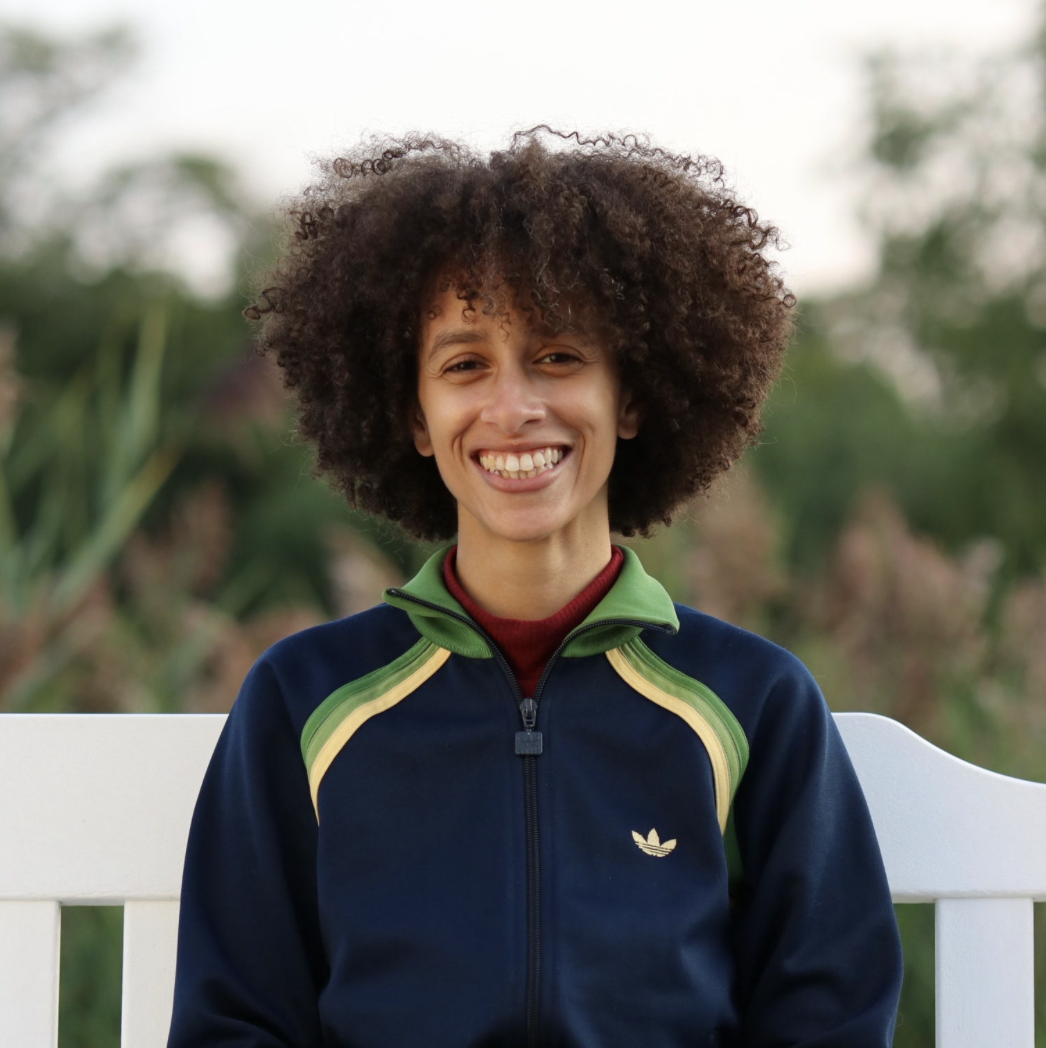 Headshot of CAF’s Researcher and Creative Consultant Joanna Preston (a mixed, Black person with curly hair) from chest up sitting on a white bench smiling in a navy jacket
