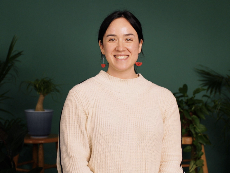 A woman with black hair and fair skin smiling, wearing red watermelon-shaped earrings and a light beige sweater, standing in front of a green background with plants to the side.