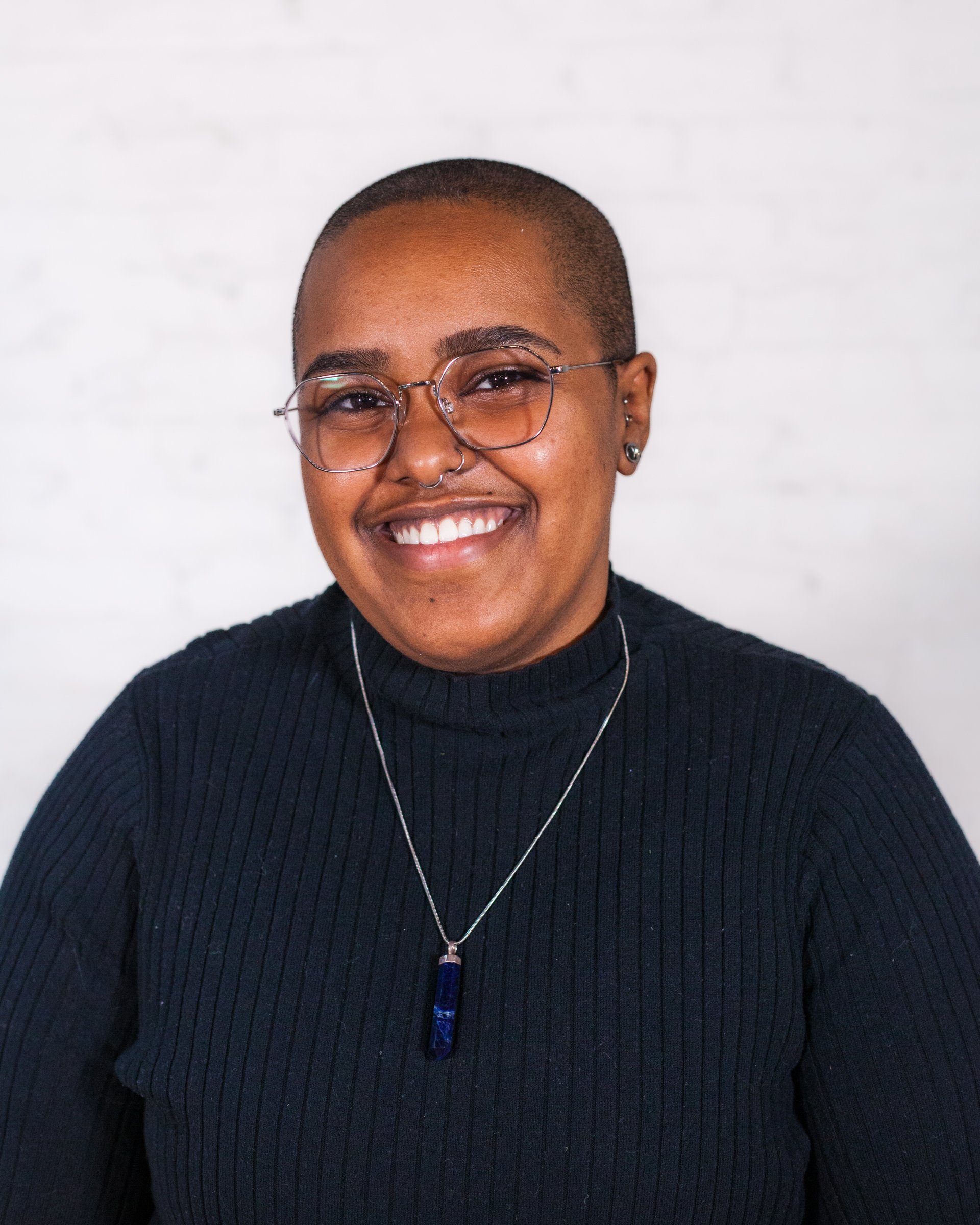Headshot of CAF’s Support Coordinator SabriAnan (a black person with a shaved head and glasses) from chest up smiling in a navy sweater and a blue pendant necklace