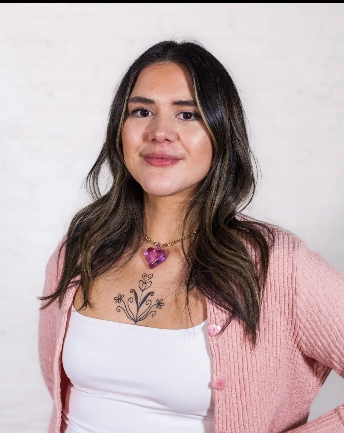 Headshot of CAF’s internal program manager Vanessa Borjon (a tan, Latine person with brown hair and a tattoo on her chest) from the chest up smiling in a white tank top, pink cardigan, and a necklace with purple pendant heart