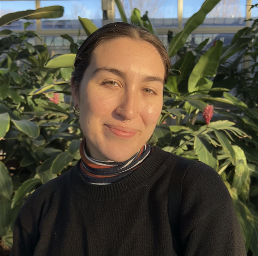 Young woman with short dark hair, wearing a light purple sweatshirt, standing in front of indoor plants and a green wall, smiling at the camera.