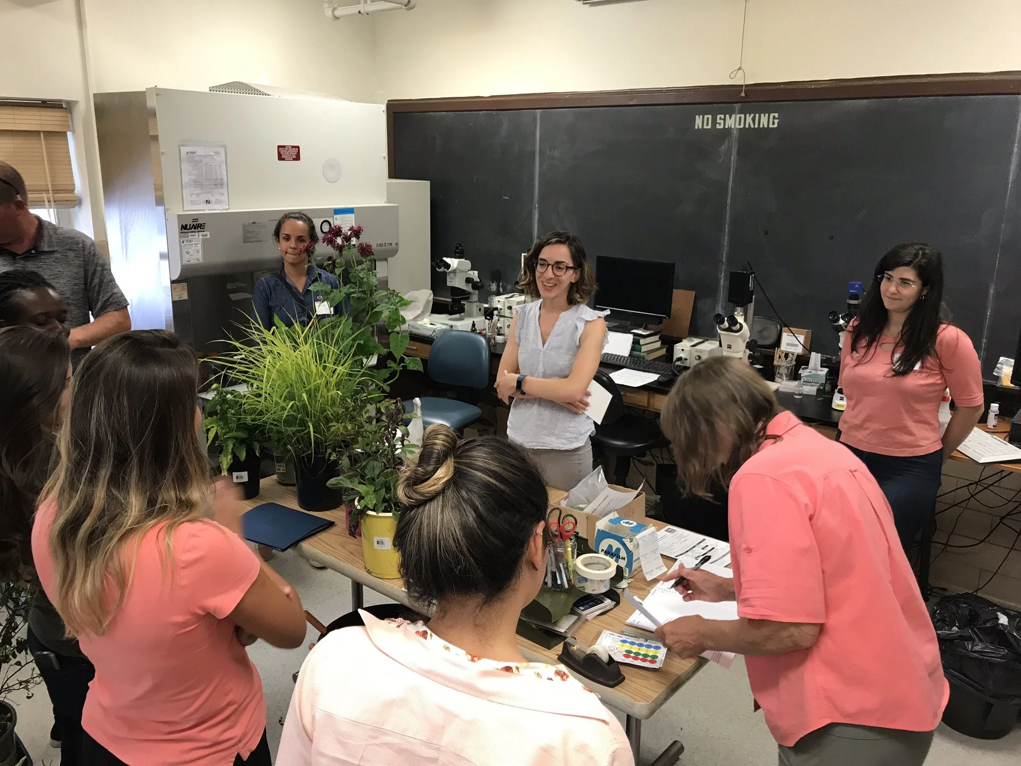 Group of women in a laboratory, with plants and scientific equipment, participating in a discussion or workshop.