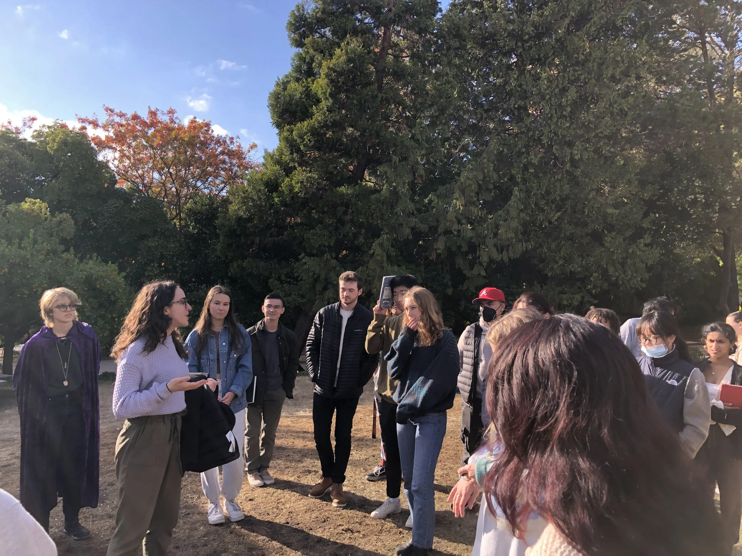 A group of young people gathered outdoors in a park, listening to a woman speaking. The group includes men and women, some wearing masks, and they are standing among trees with the sun shining through.