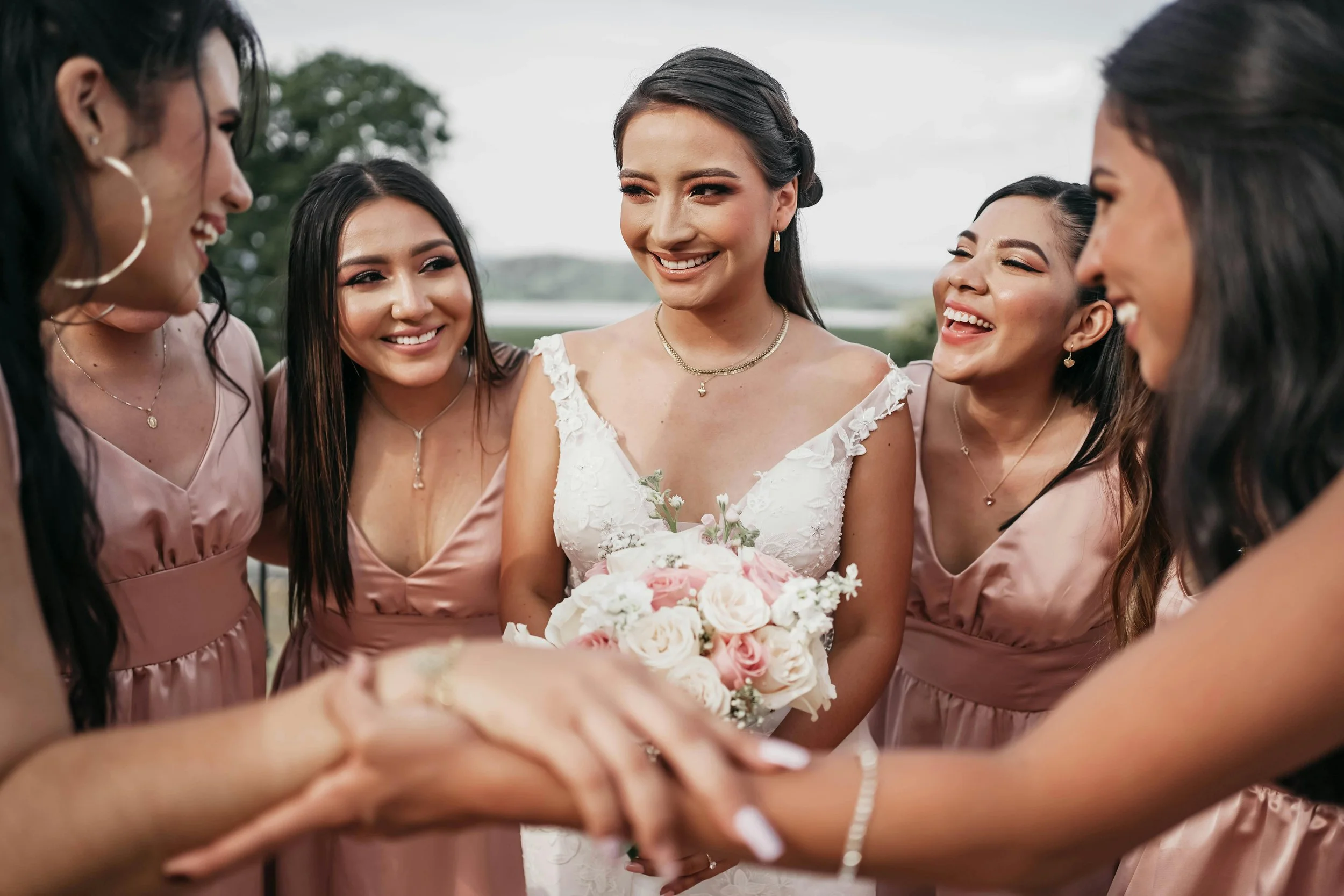A bride in a white lace wedding dress holding a bouquet of pink and white roses, smiling with her bridesmaids in matching rose-colored dresses, all joyful and laughing together outdoors.