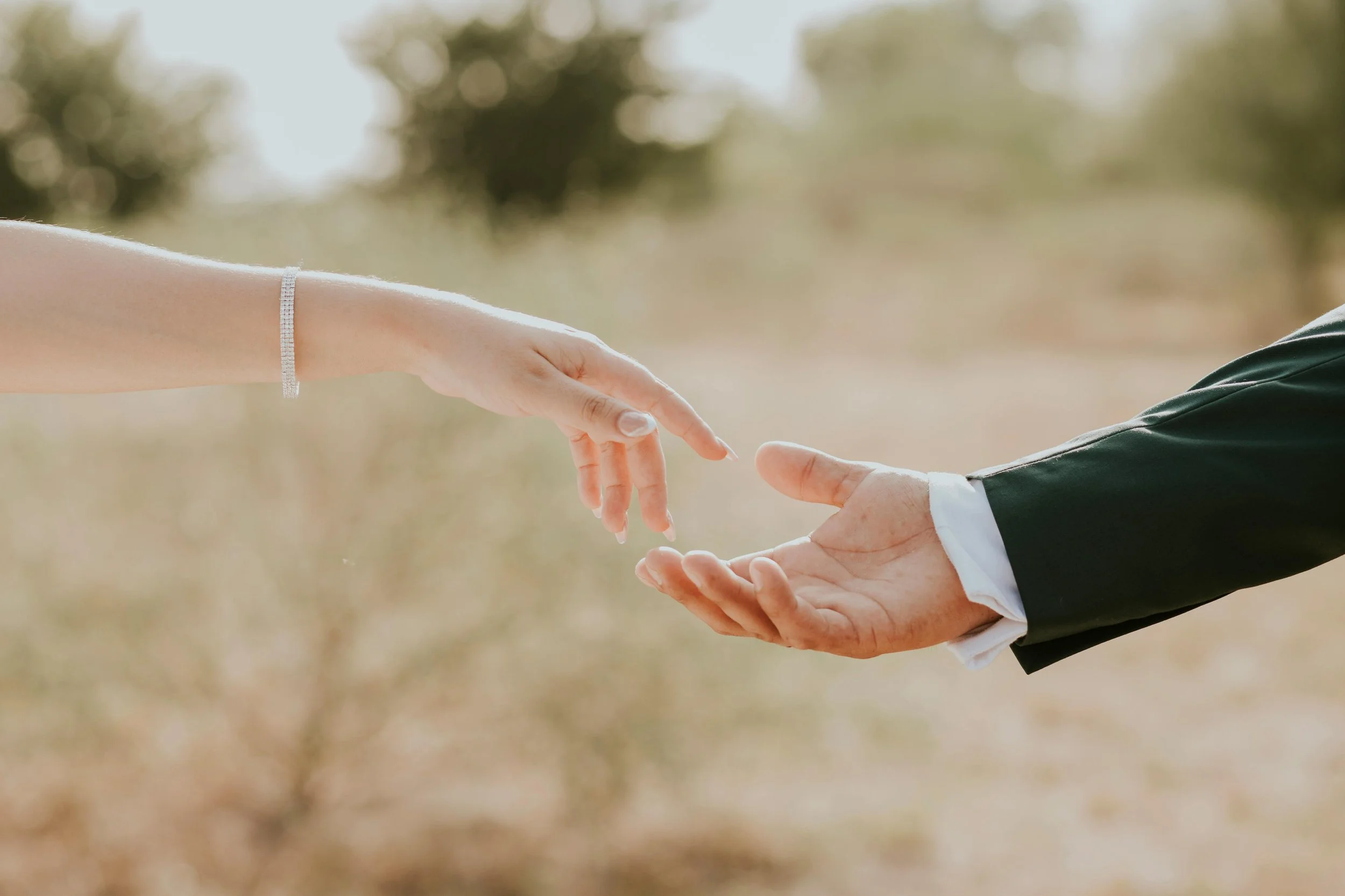 A woman reaching out her hand to touch a man's hand, outdoors with a blurred natural background.