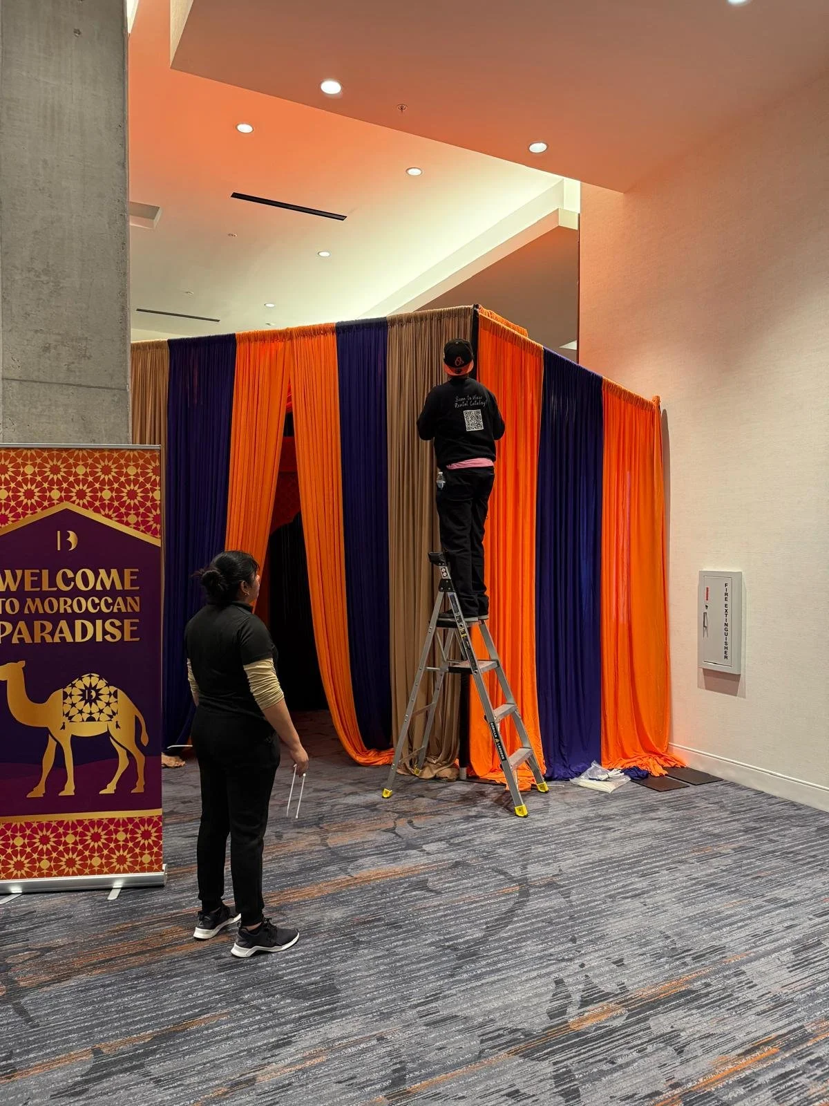 Two workers are setting up colorful fabrics, orange, purple, and tan, around an entrance to a Moroccan-themed event called 'Moroccan Paradise'. One worker is on a ladder adjusting the fabric, while the other stands nearby. There's a welcome sign with