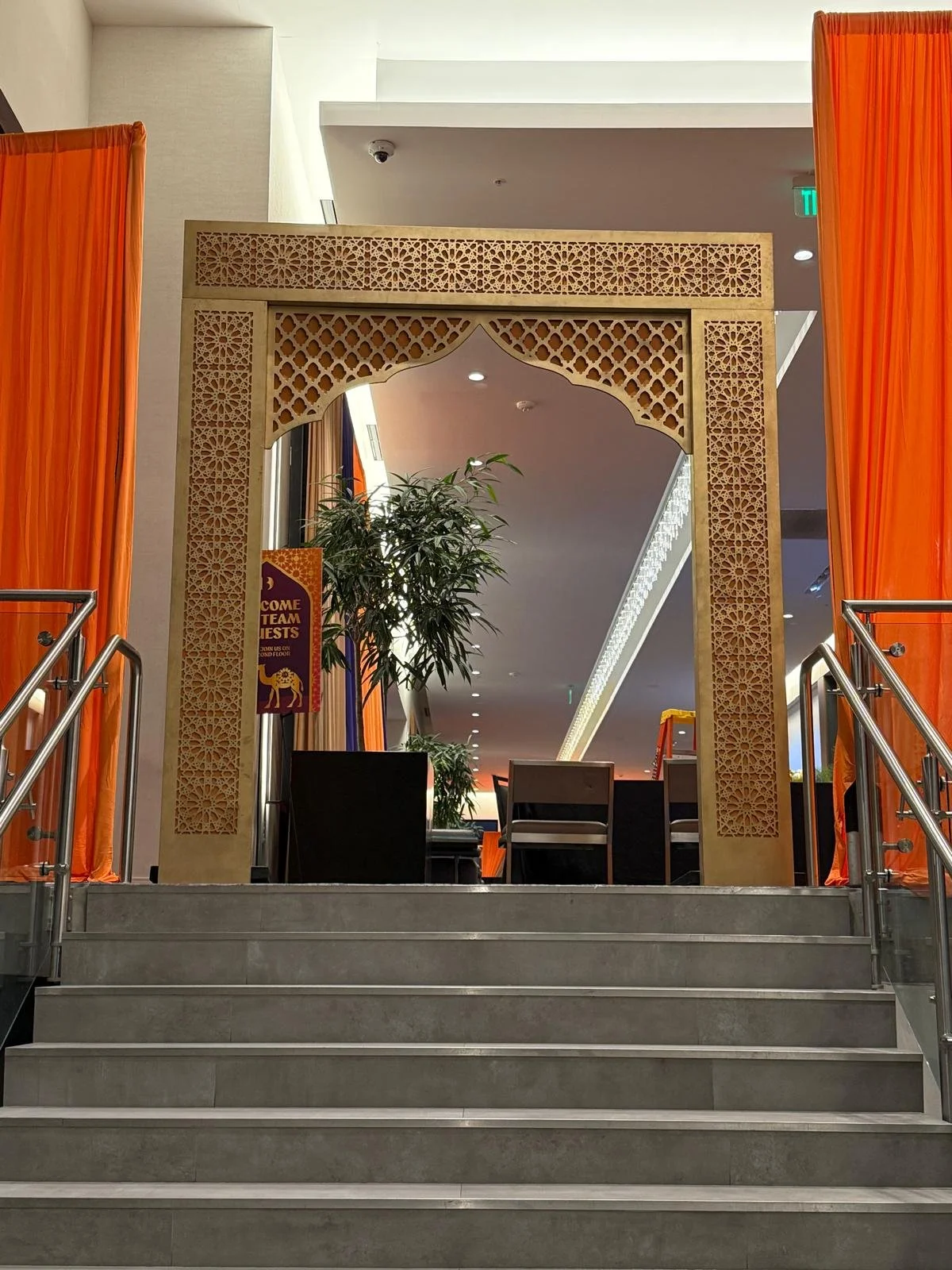 An indoor area with stairs leading up to a decorative archway with intricate patterns, orange curtains on either side, and potted plants inside.