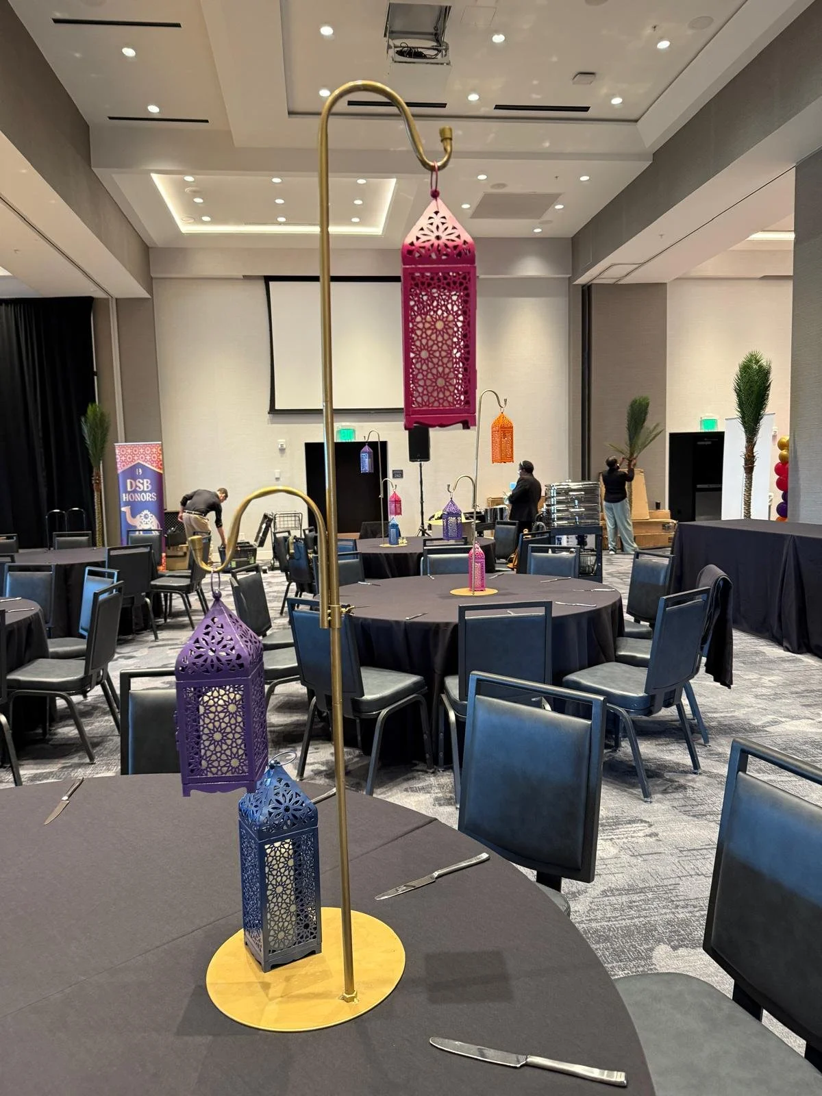 Event space with round tables covered in black tablecloths, decorated with colorful lantern centerpieces. The room has gray walls, a ceiling with recessed lighting, and a few people preparing or setting up. There is a screen or projector at the front