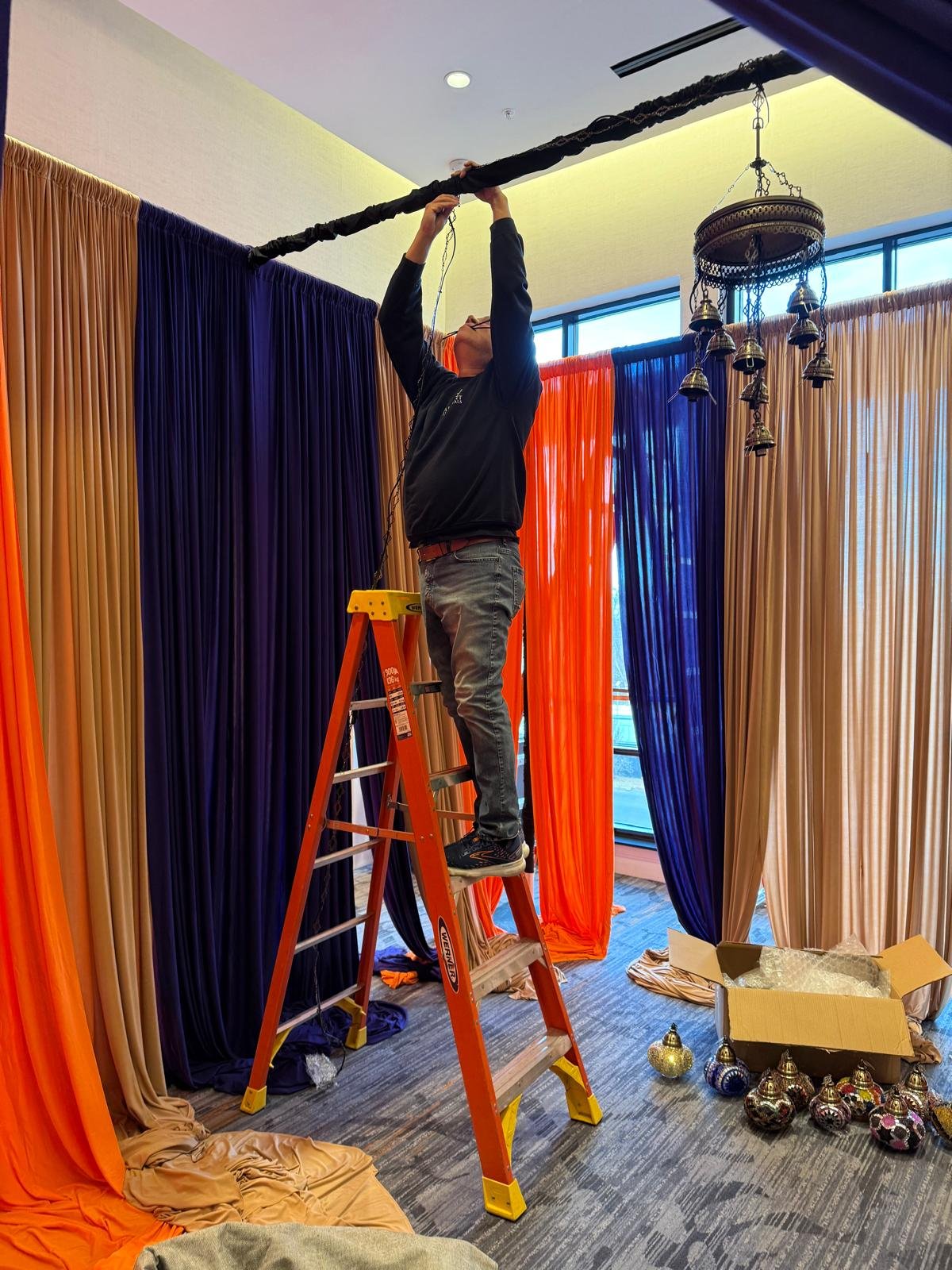 A person standing on an orange ladder adjusting a black curtain rod near a chandelier with multiple hanging bells, in a room with colorful curtains and a cardboard box filled with decorative ornaments.