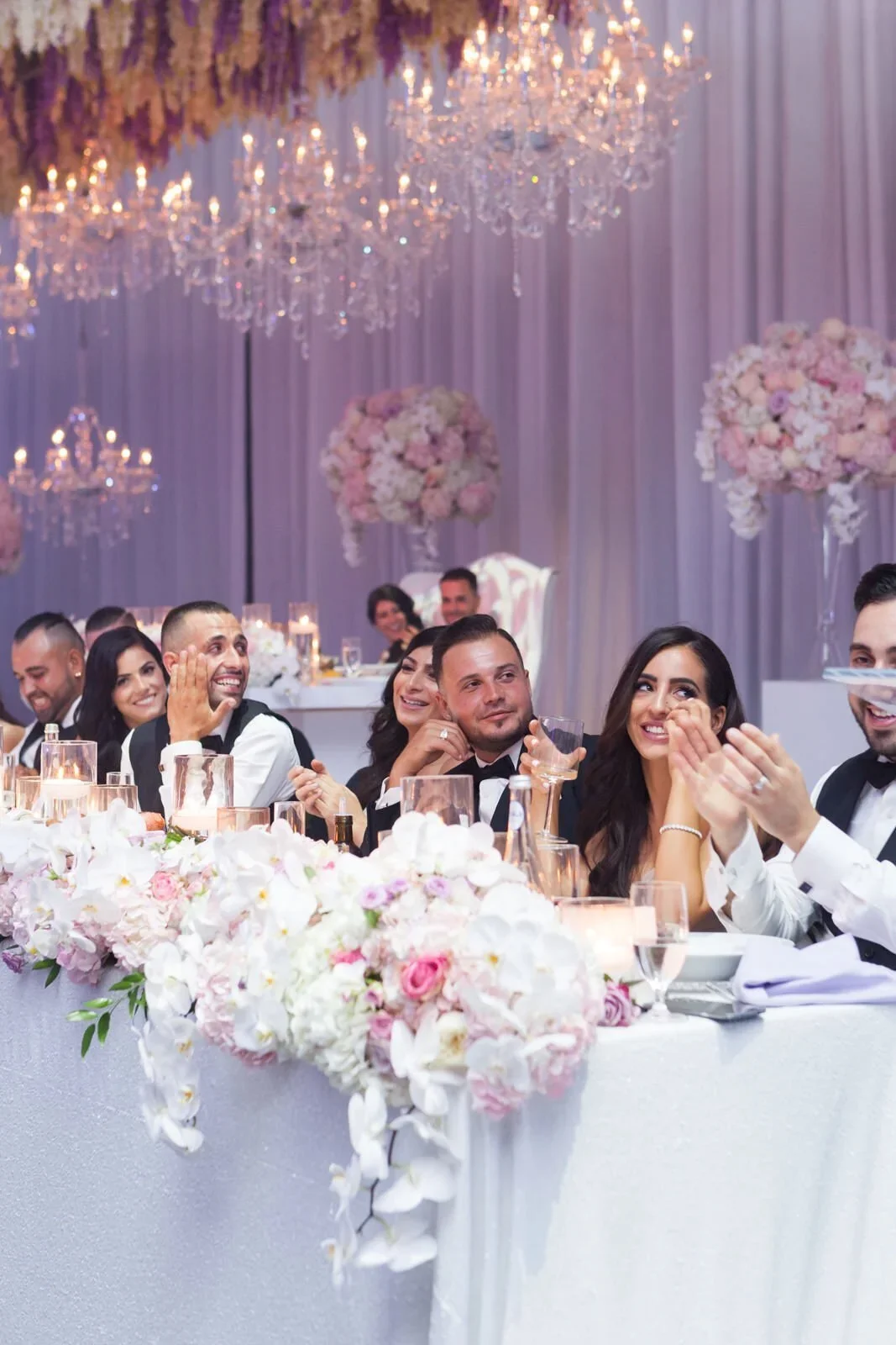 Group of wedding guests sitting at a decorated reception table, smiling and clapping, with chandeliers and floral arrangements overhead.