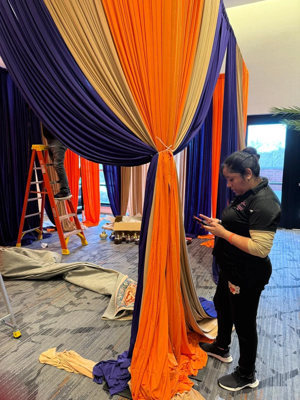 Interior of a room with orange, purple, and beige drapes being installed or arranged, with a woman looking at her phone and a person on a ladder in the background.