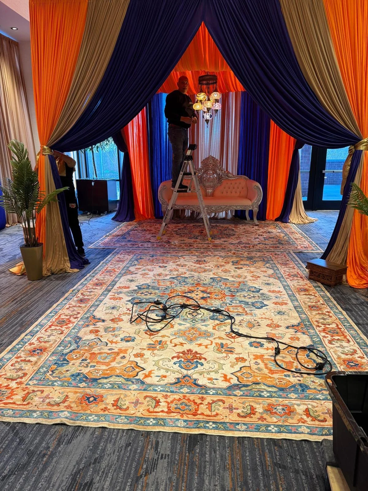 A decorative event backdrop with blue, orange, gold, and beige drapes hung around a stage area, with a vintage-style cream and gold upholstered seating piece. Two large potted plants are on either side, and a patterned area rug covers the floor. Two 