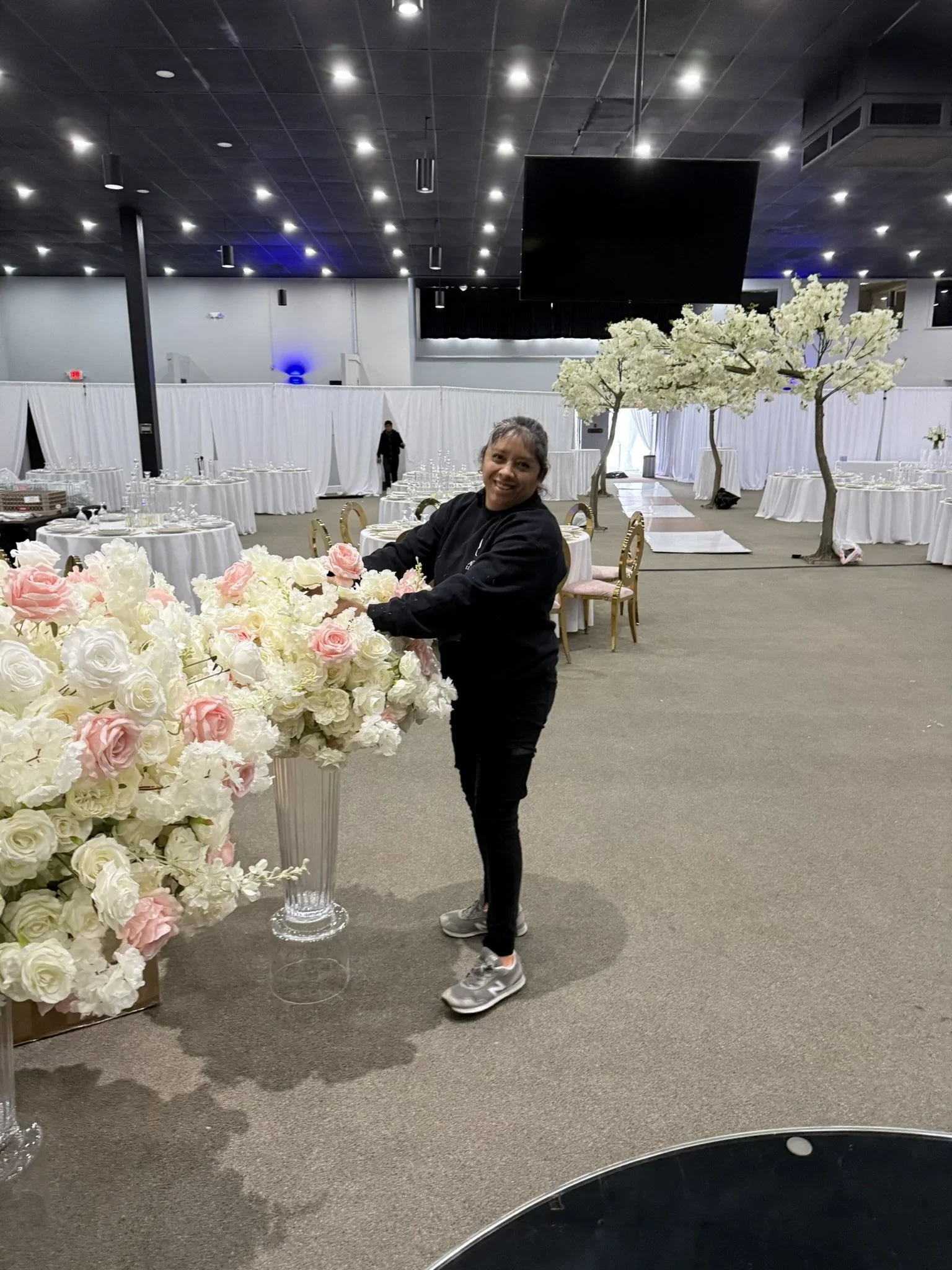 Woman arranging pink and white flowers in a large glass vase in a banquet hall decorated with white tables, pink and white floral arrangements, and white curtains, preparing for an event.
