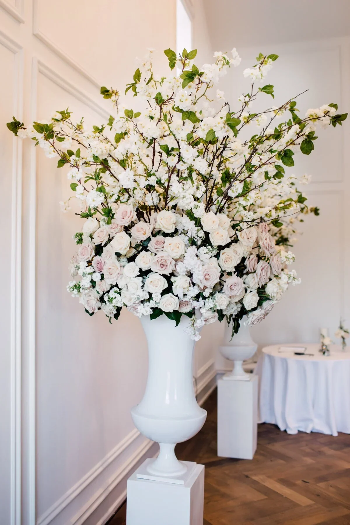 Large white floral arrangement with roses and branches in a tall white vase, on a pedestal in a decorated room.