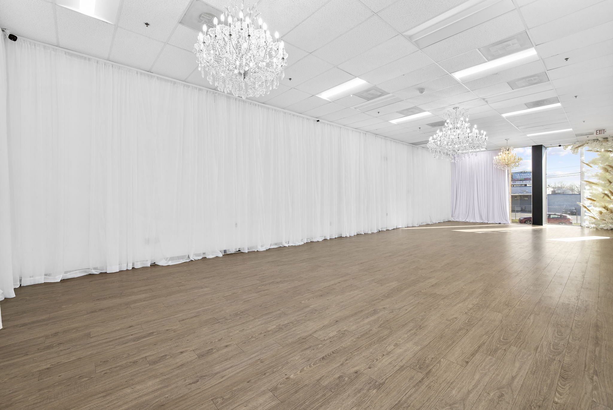 Spacious empty room with wooden floor, white walls, ceiling chandeliers, and large windows.