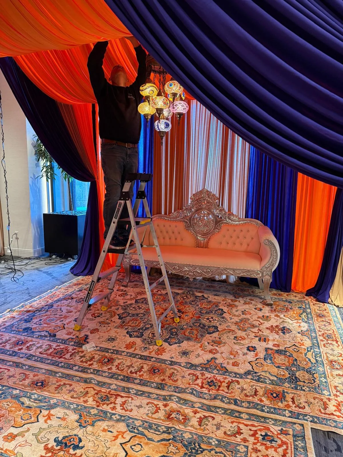A person standing on a ladder adjusting colorful hanging lanterns under orange, purple, and blue drapes in a decorated room with a vintage sofa, patterned rug, and potted plant in the background.