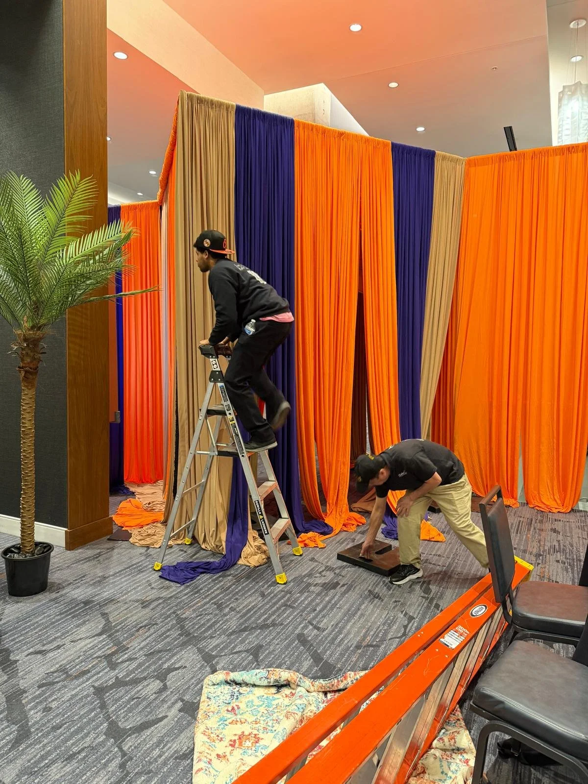 Two workers are setting up colorful orange, blue, and beige curtains in an interior space. One is standing on a ladder, while the other is crouching on the floor adjusting the fabric.