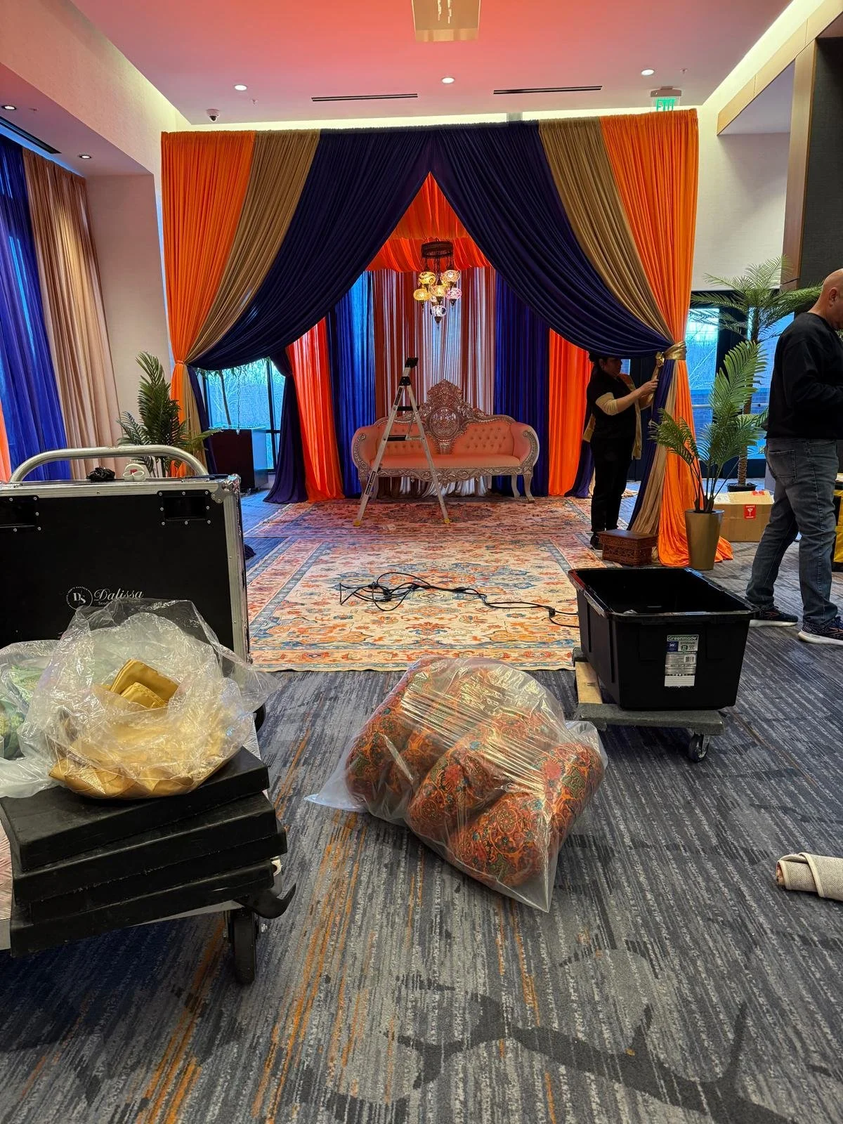 A decorated stage with orange, navy blue, and gold curtains, a vintage-style pink sofa, chandelier, and a patterned rug, with people setting up and supplies around.