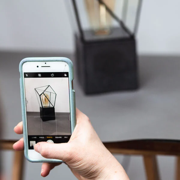 A hand holding a smartphone to photograph a small handmade product in natural light.
