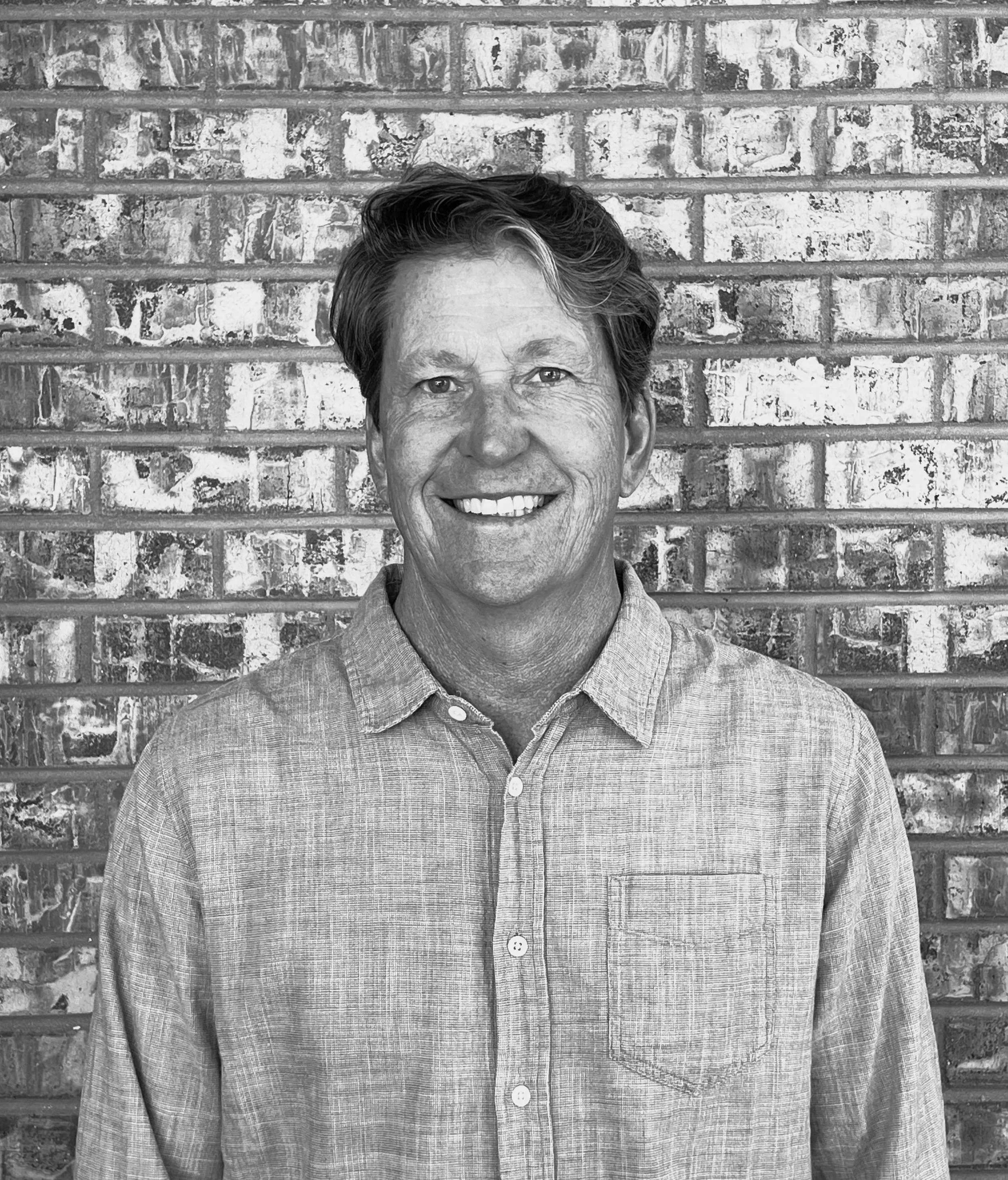 Black and white photo of a smiling middle-aged man with short, slightly wavy hair, wearing a collared, button-up shirt, standing against a textured brick wall.