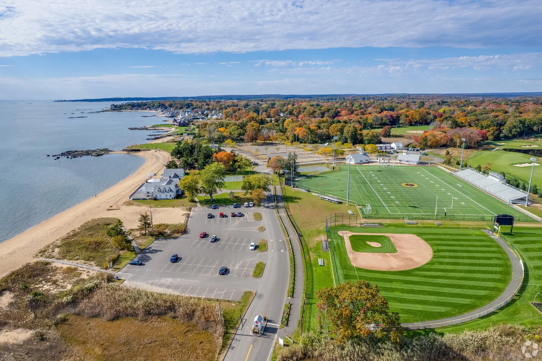 Aerial view of a coastal park with a beach, parking lots, a football field, and a tree-lined area with autumn foliage.