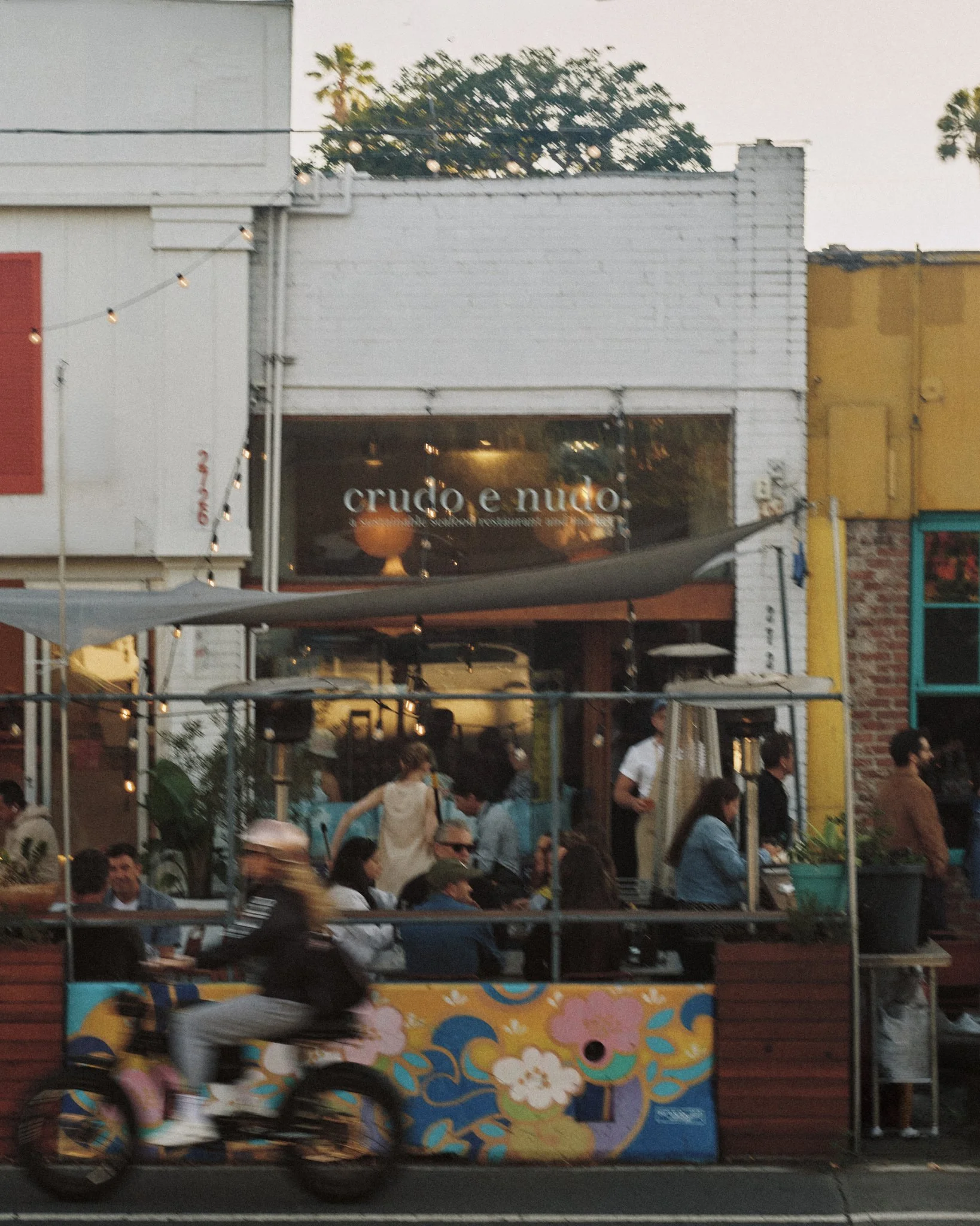 Street view of Crudo e Nudo with guests dining on the patio and a bicyclist riding by