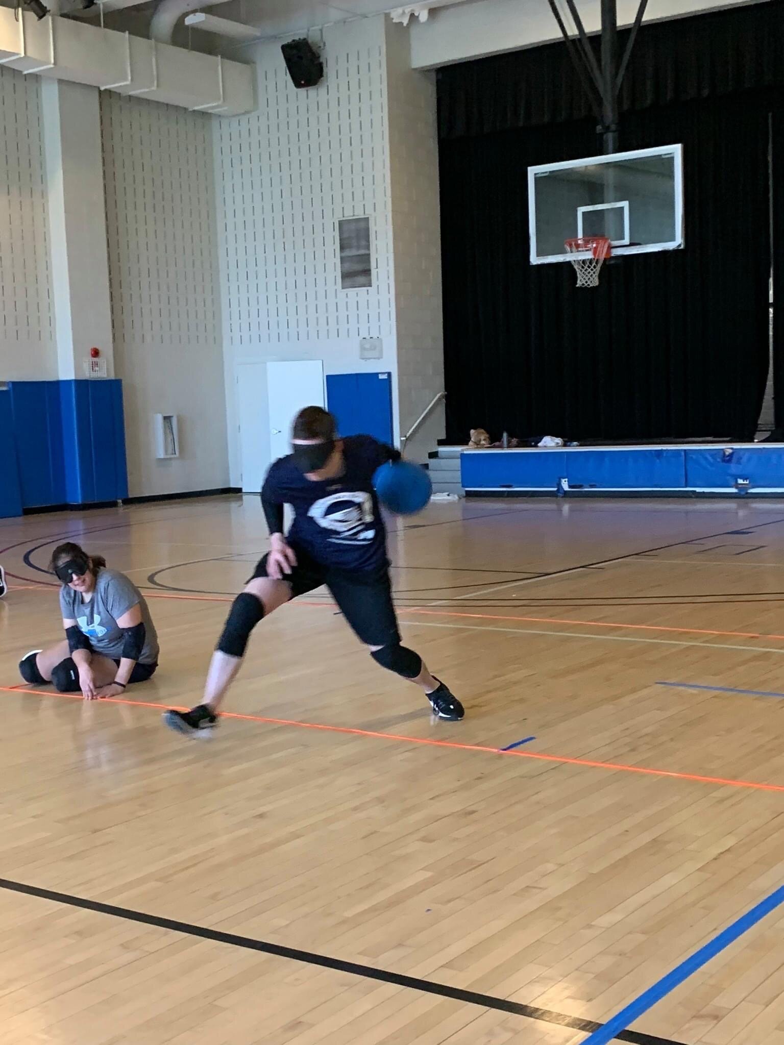 Action shot: WMABA Goalball player Caleb Berkemeier in the midst of throwing, and Lori, another player, is laying on the court in defensive position.
