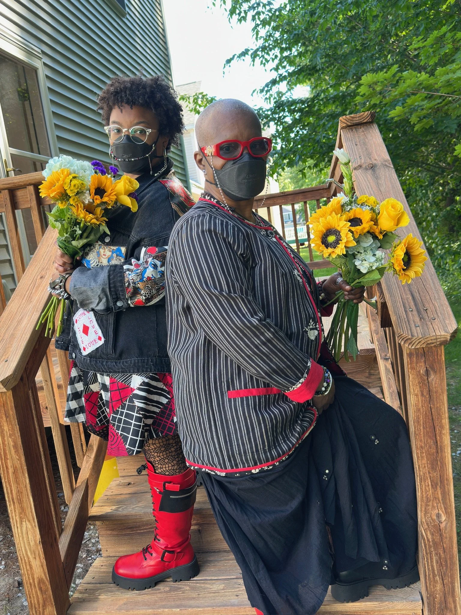 A photo of Amaranthia (a younger Black woman with an afro) and Claire (older Black woman with a shaved head) both wearing black N95s and glasses. Both are wearing black and red themed alternative style outfits, holding sunflower bouquets on a porch.