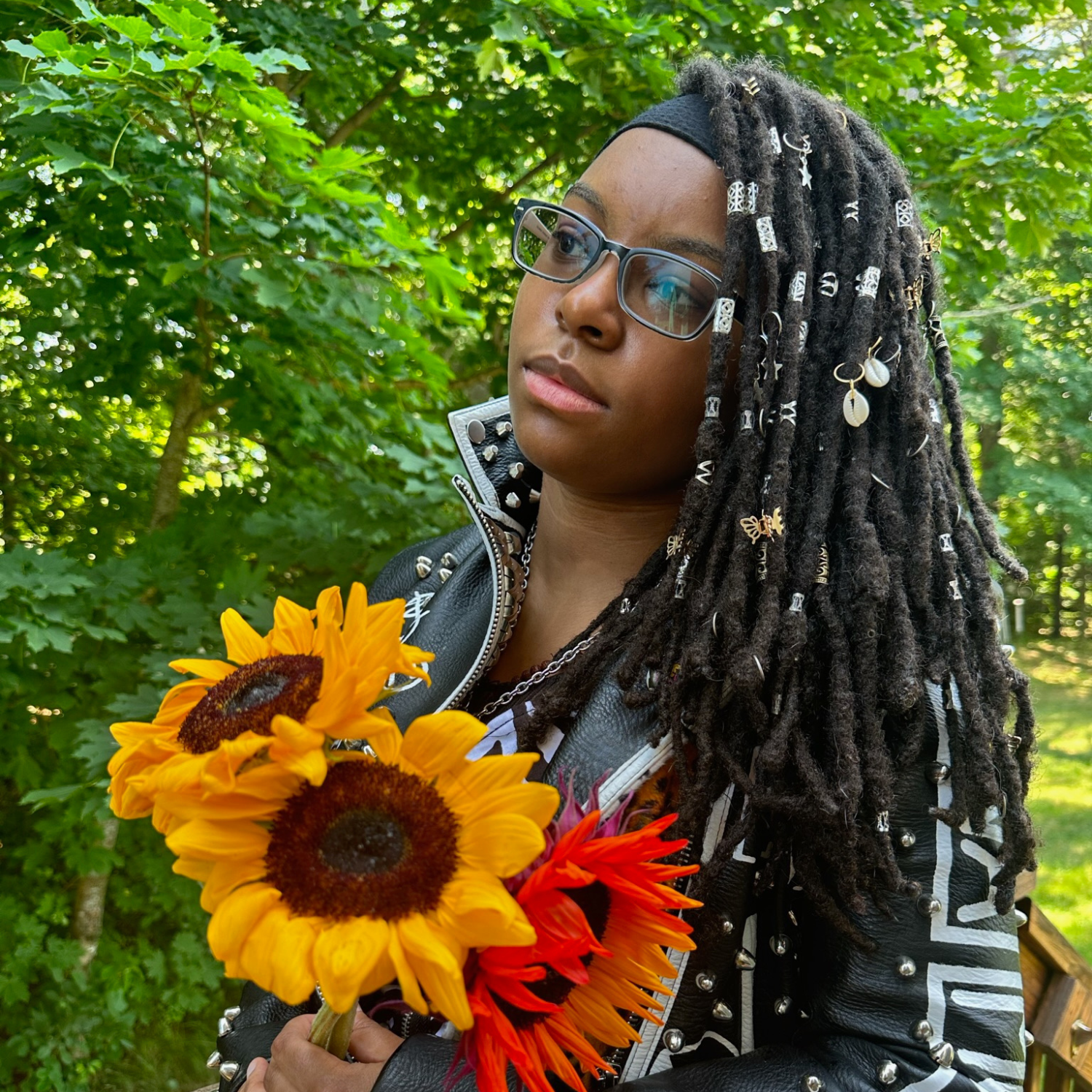 Amaranthia is a Black woman with dreadlocks. She's turned to the left with a neutral expression while wearing black rimmed glasses. Amaranthia is wearing a black leather jacket and holding three sunflowers. Bushy trees are behind her.