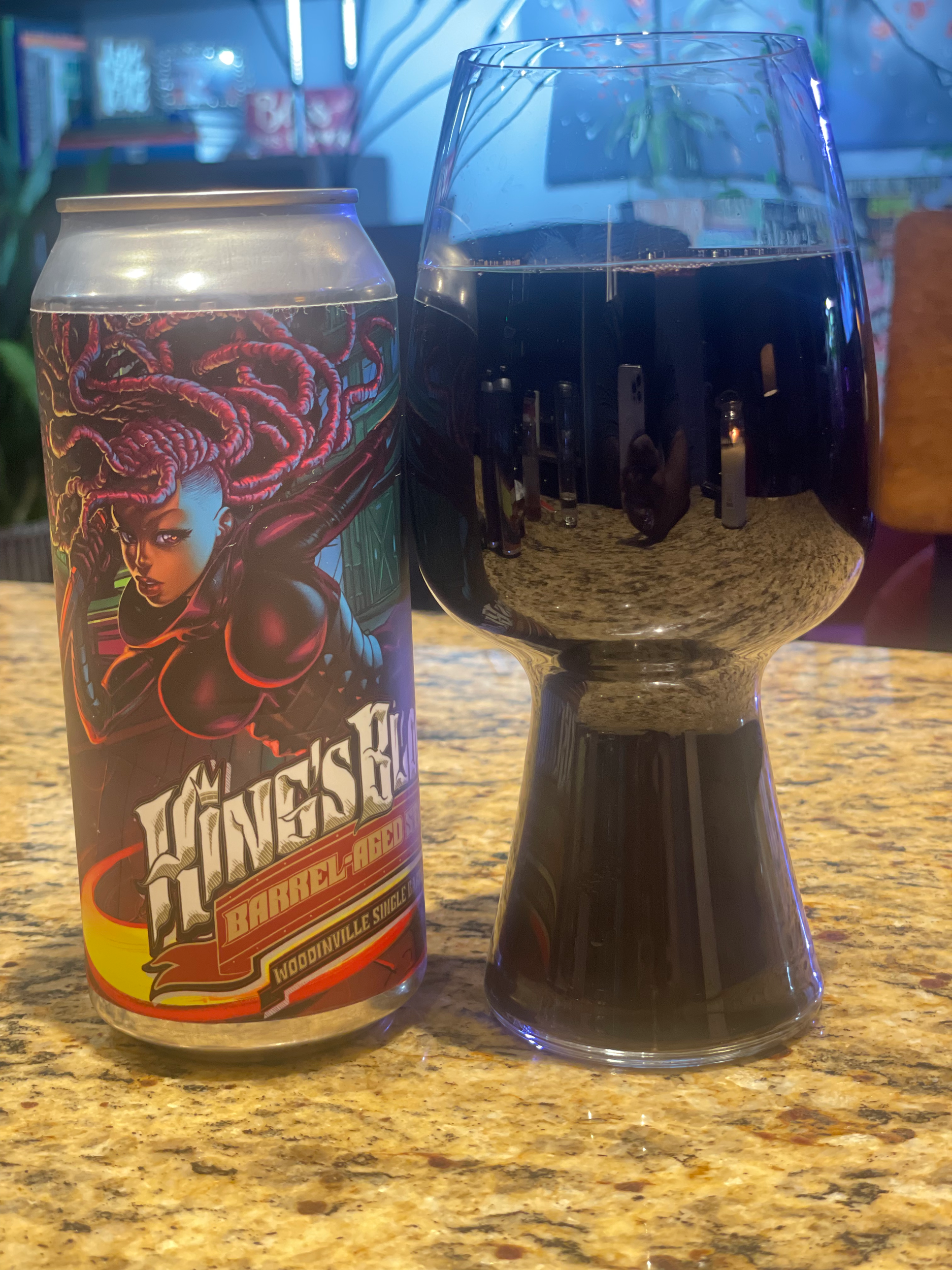 A can of Warstic baseball beer with vibrant artwork of a woman with dreadlocks, and a tall glass filled with dark beer, both placed on a granite countertop.