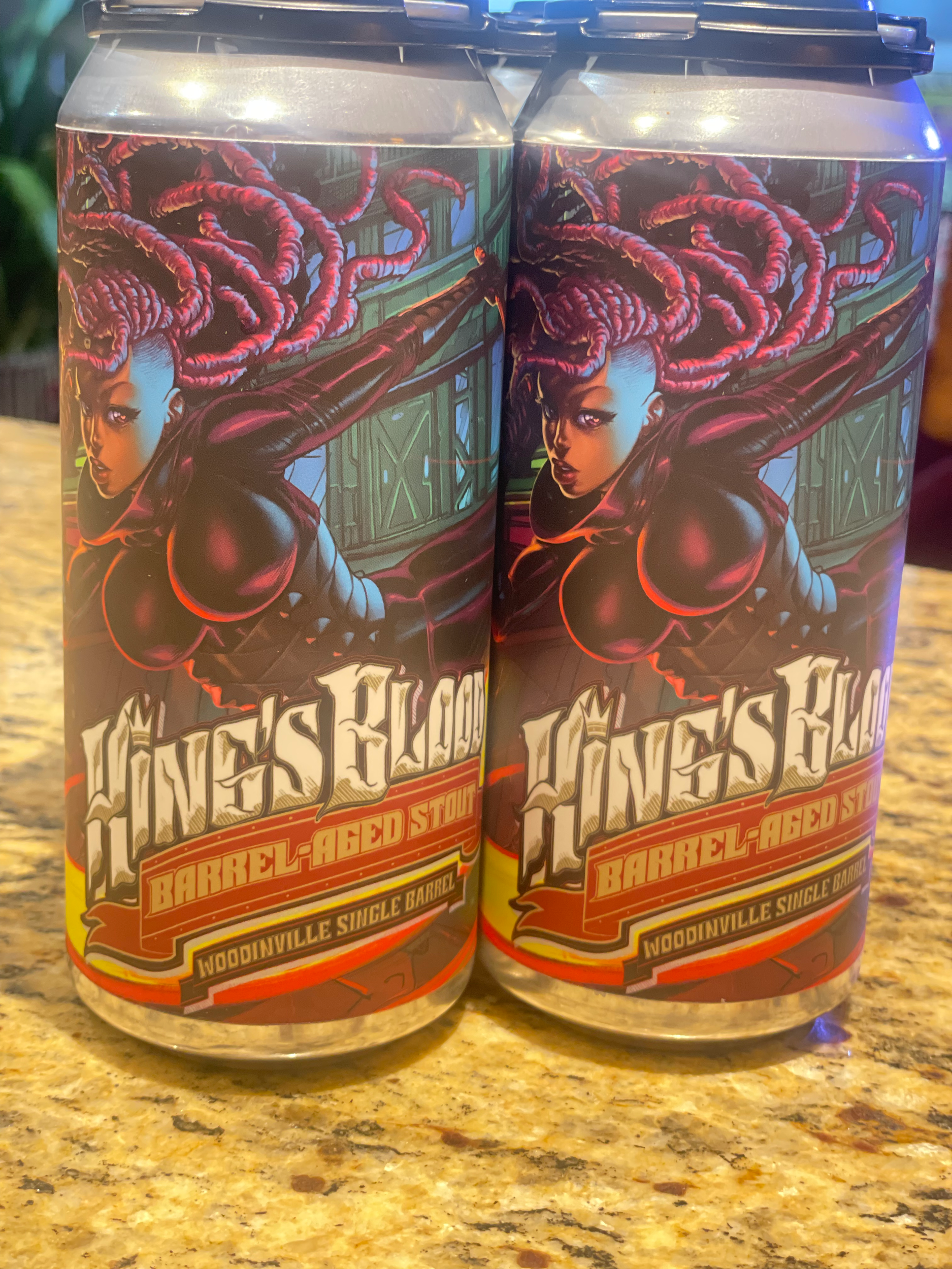 Two cans of beer with colorful superhero-themed labels featuring a female character with purple dreadlocks and a black bodysuit, with the "Barrel-Aged Stout" on the labels.