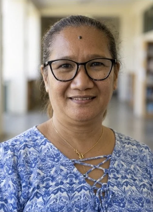 A woman with glasses and a gold necklace, smiling, in a well-lit indoor setting with bookshelves in the background.